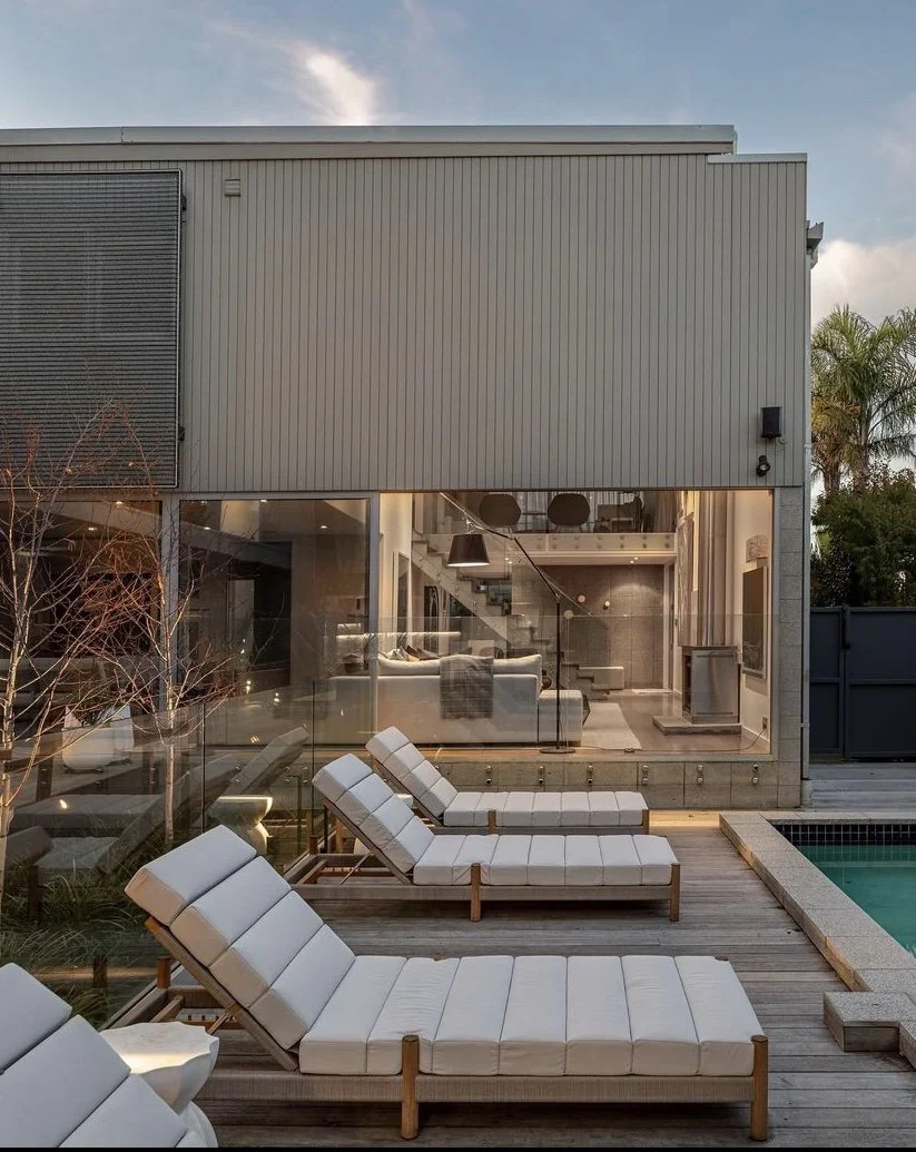 Modern architectural home exterior with a lounge area featuring white cushioned sunbeds on a wooden deck next to a swimming pool. The house has large glass windows revealing a contemporary interior living space.