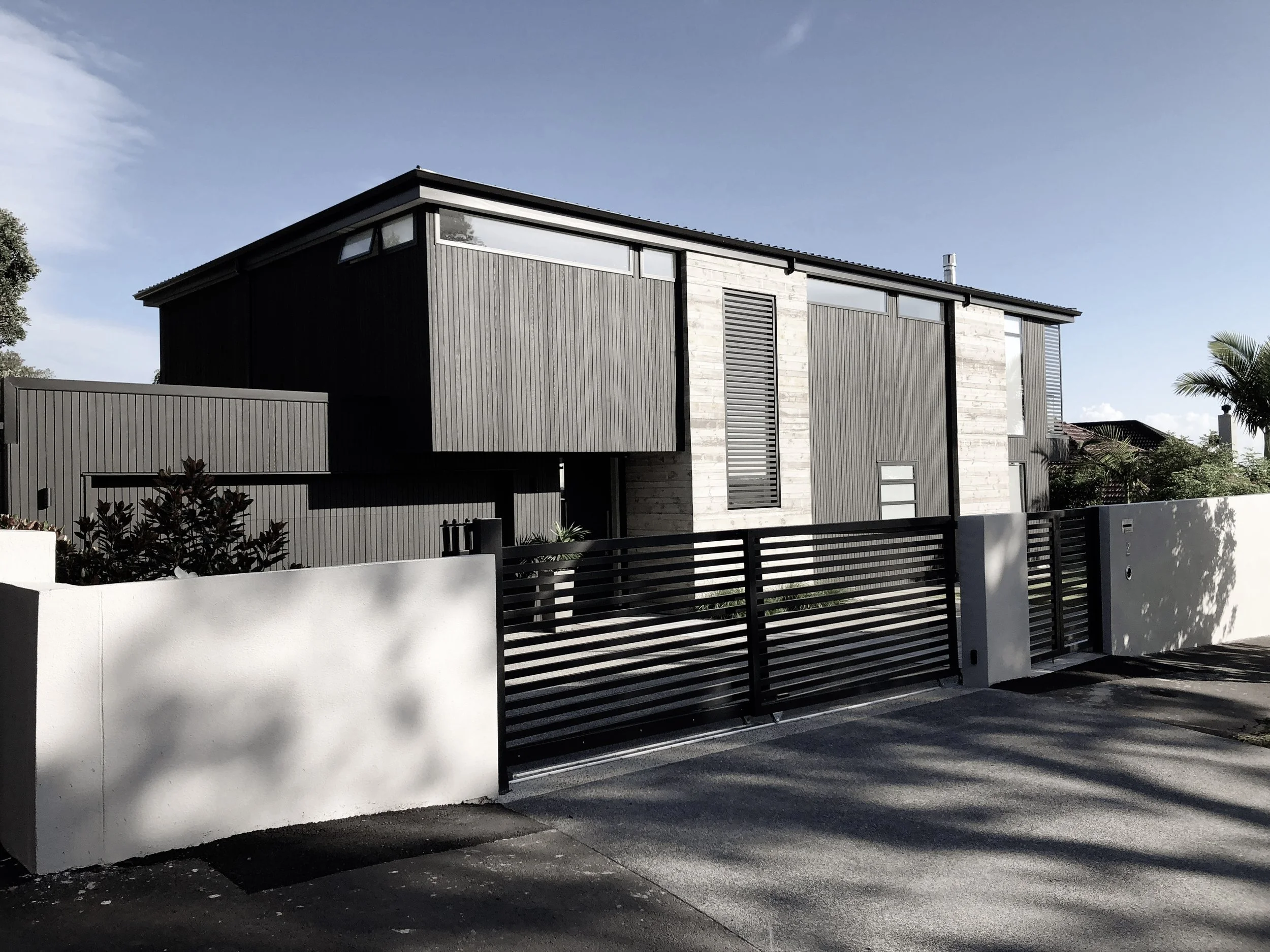 Modern two-story house with dark vertical wooden siding, large windows, and a prominent white brick accent wall, surrounded by a white fence and a black gate, under a clear blue sky.