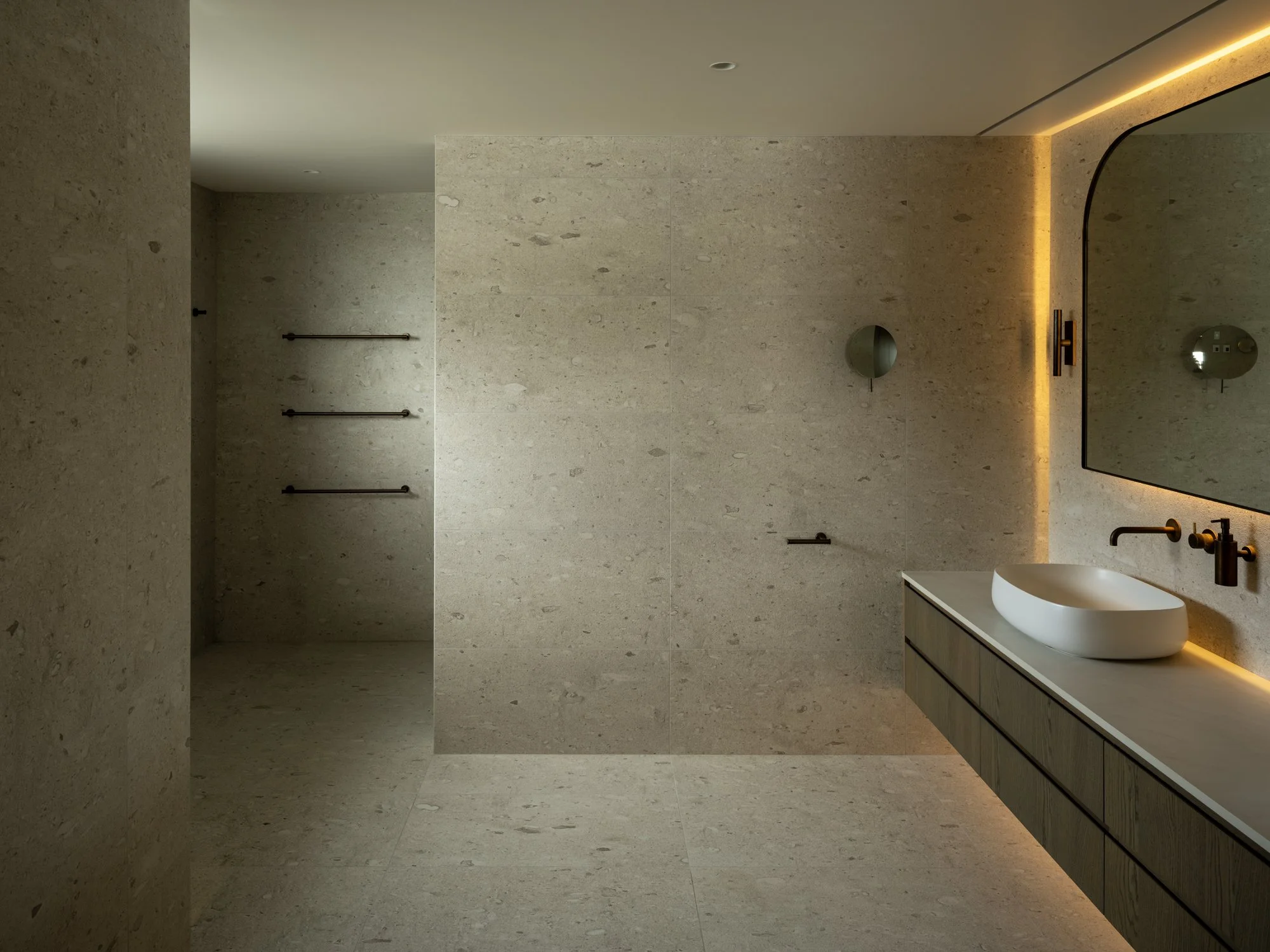 Modern bathroom with beige stone walls and floor, black fixtures, a mirror with warm lighting, a sink on a wooden vanity, and minimal decor.