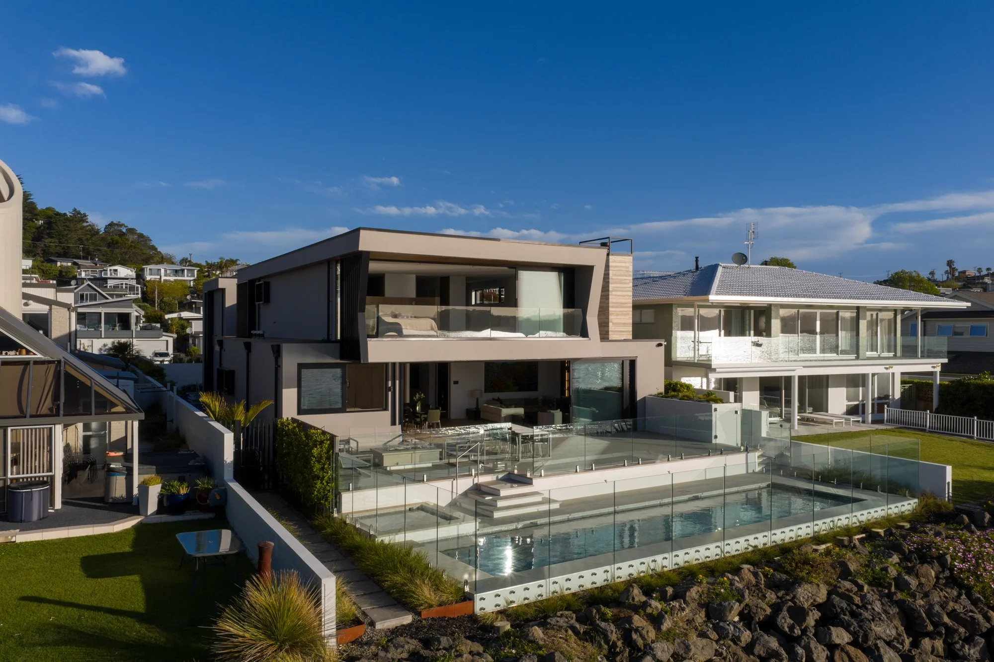 Modern multi-story architectural home with large glass windows, a swimming pool, and a landscaped garden in a coastal area overlooking the ocean under a blue sky.