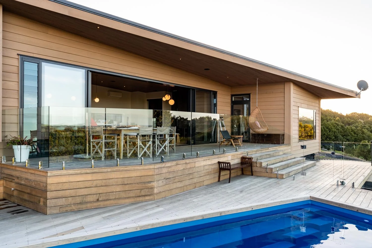 Modern house with wooden exterior, glass balcony, outdoor dining area, hanging swing chair, and swimming pool at sunset.