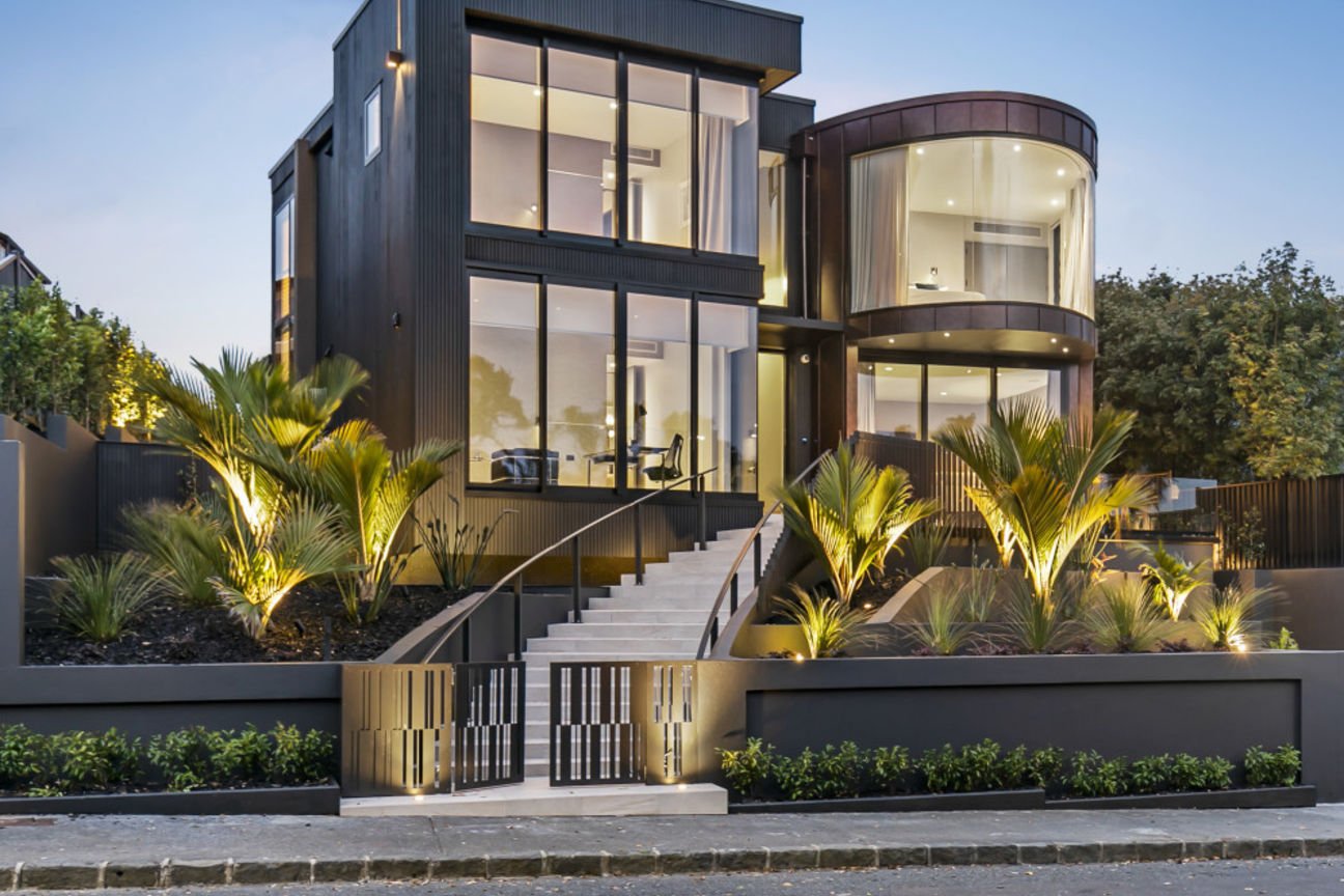 Architecturally designed custom modern black house with large glass windows, illuminated outdoor landscaping, and a staircase leading to the entrance.