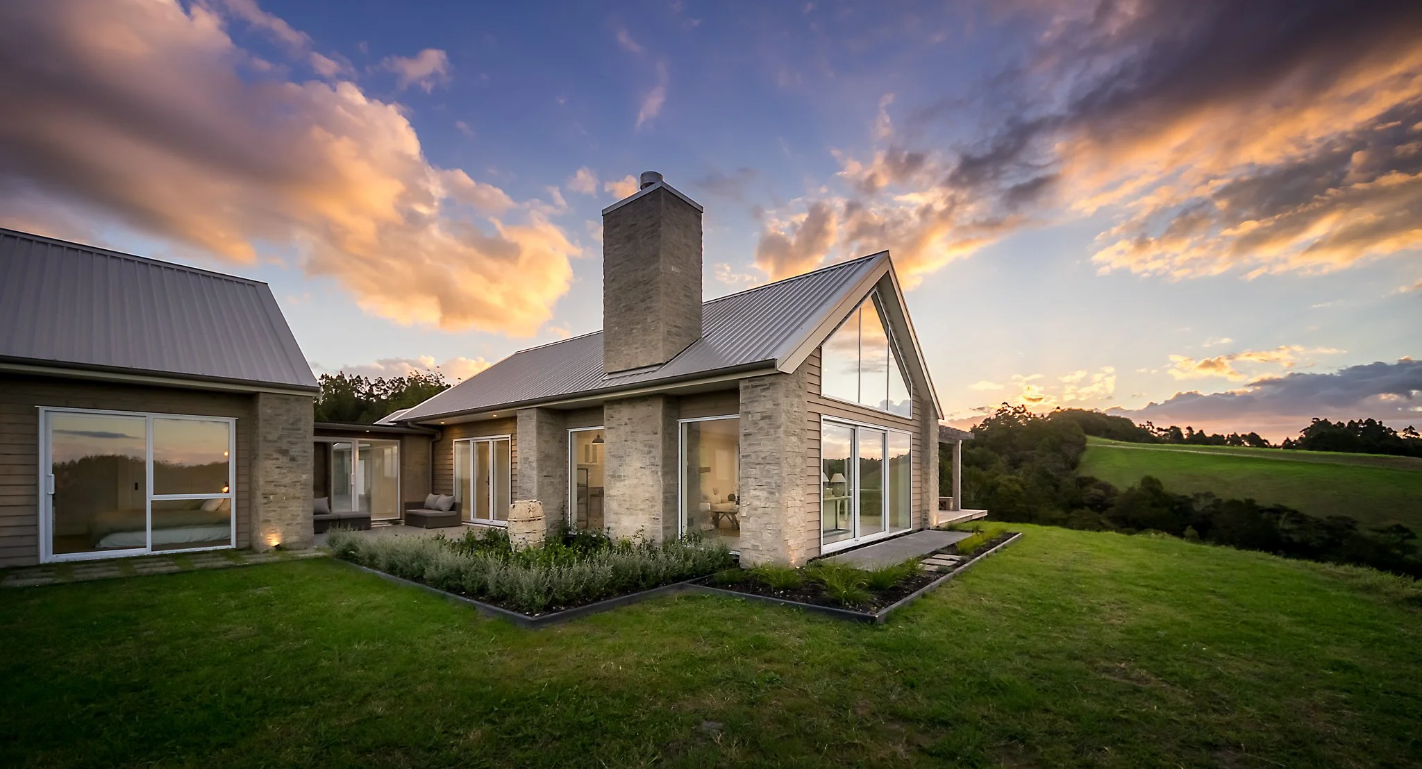 Custom rural architectural home with large windows and a stone chimney, set on a grassy country landscape during sunset.