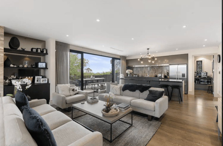 Living room with white sofas, black pillows, a coffee table with books and a plant, open kitchen with island and bar stools, sliding glass door leading to outdoor patio, modern decor.