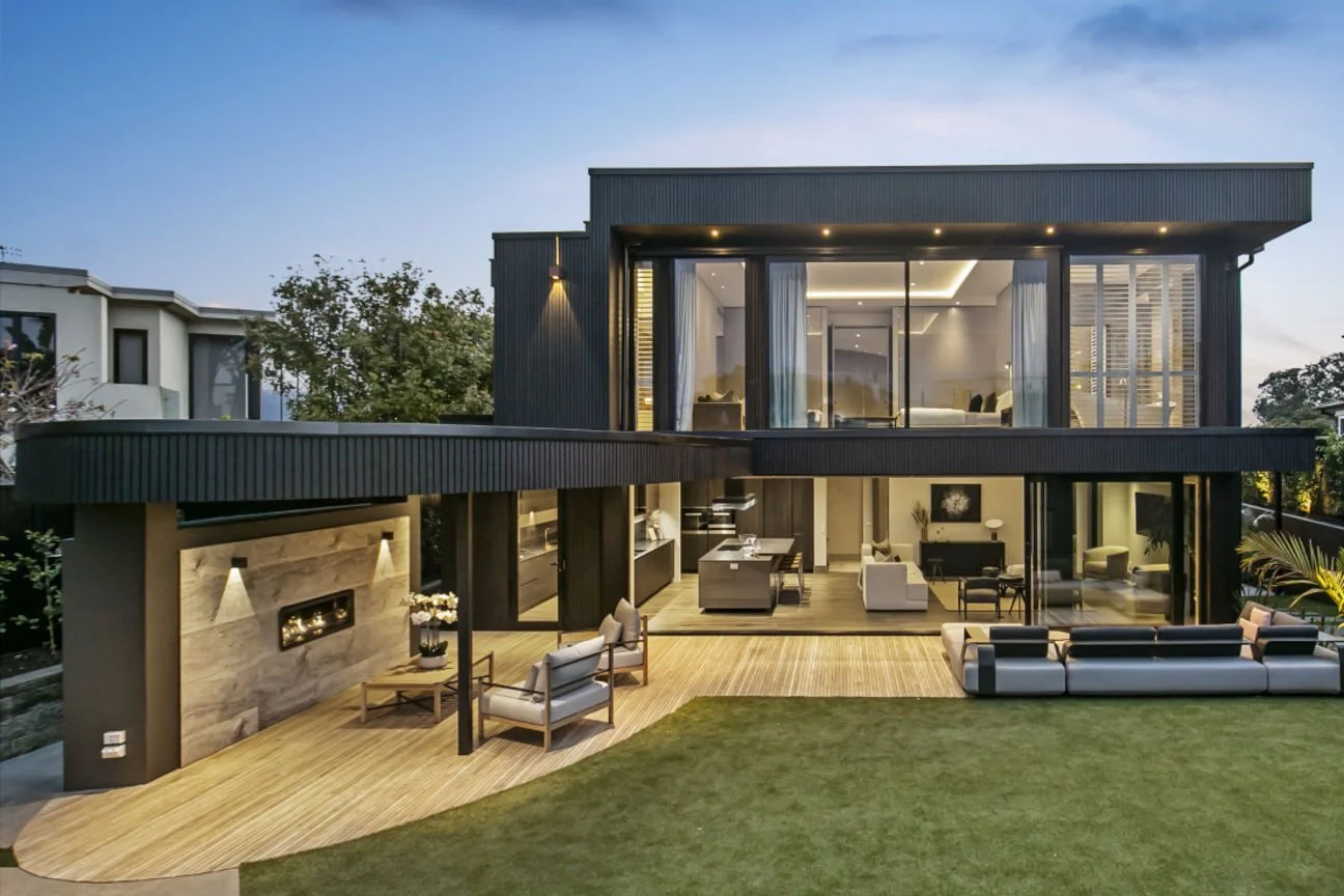 Modern multi-story newbuild custom home with large glass windows, outdoor patio with seating, and a well-maintained lawn at dusk.