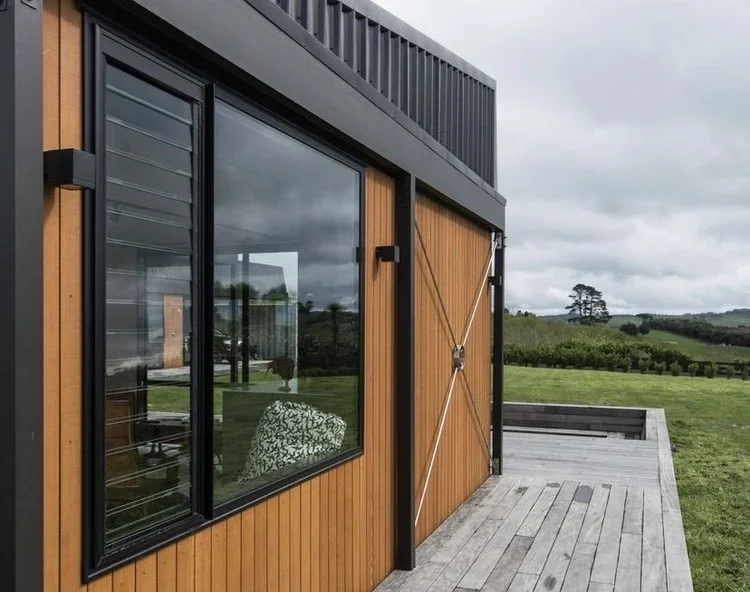 Close-up of a modern house with wooden siding, large black-framed sliding glass door, and a wooden deck, with a grassy landscape and cloudy sky in the background.