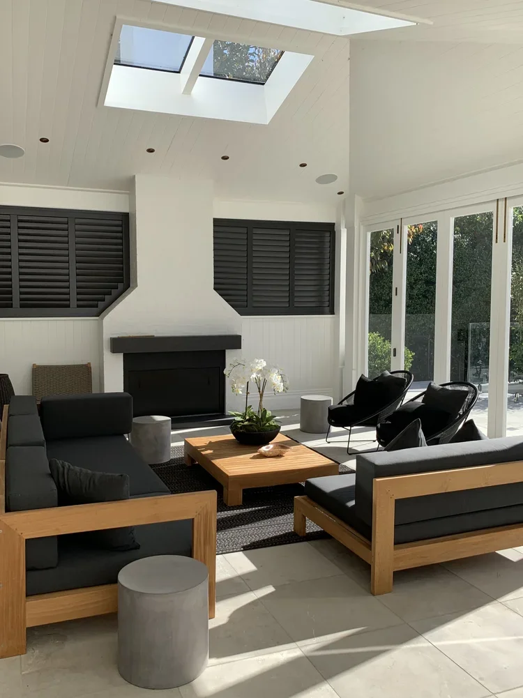 Bright living room with skylight, white walls, black window shutters, and modern black and wood furniture, including sofas, chairs, and a coffee table with a potted orchid.