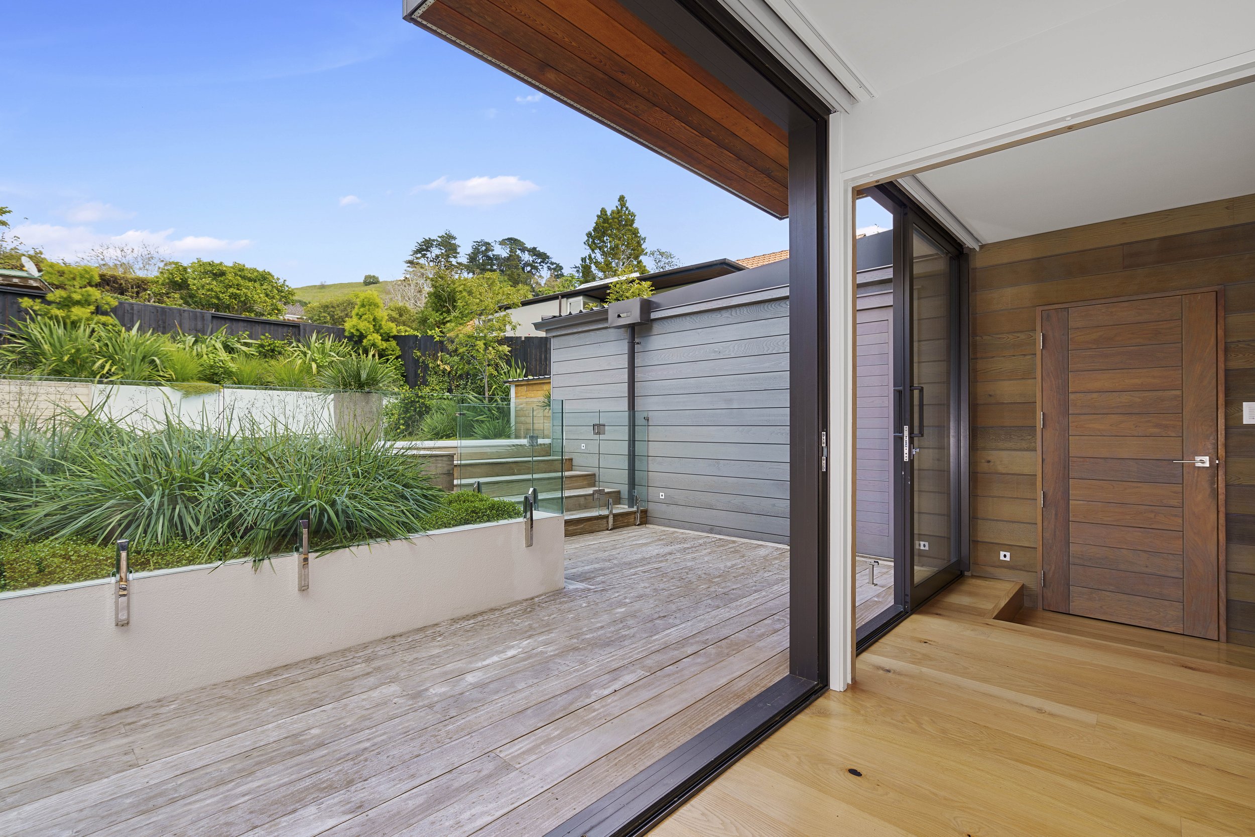 View of a modern outdoor patio area with wooden flooring, glass railing, and lush greenery beyond. The patio opening features sliding glass doors with wooden interior finish and a ceiling with wooden paneling, overlooking a landscaped backyard with p