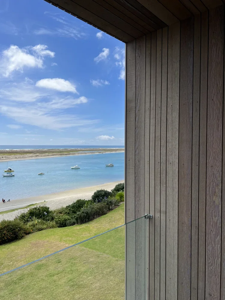 View of a beach and water from a window with wooden walls, boats floating on the water, green grass, and blue sky with clouds.