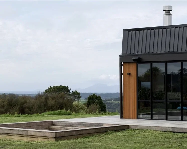 Part of a modern house with large glass windows, a wooden exterior, and a metal roof, set in a grassy landscape with trees and distant mountains.
