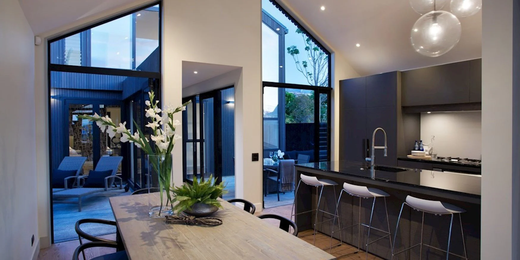 Modern kitchen with black cabinets, gray walls, a black countertop with a sink, three white barstools, and a dining table with a vase of white flowers; large windows and sliding doors reveal an outdoor patio with lounge chairs and outdoor seating.