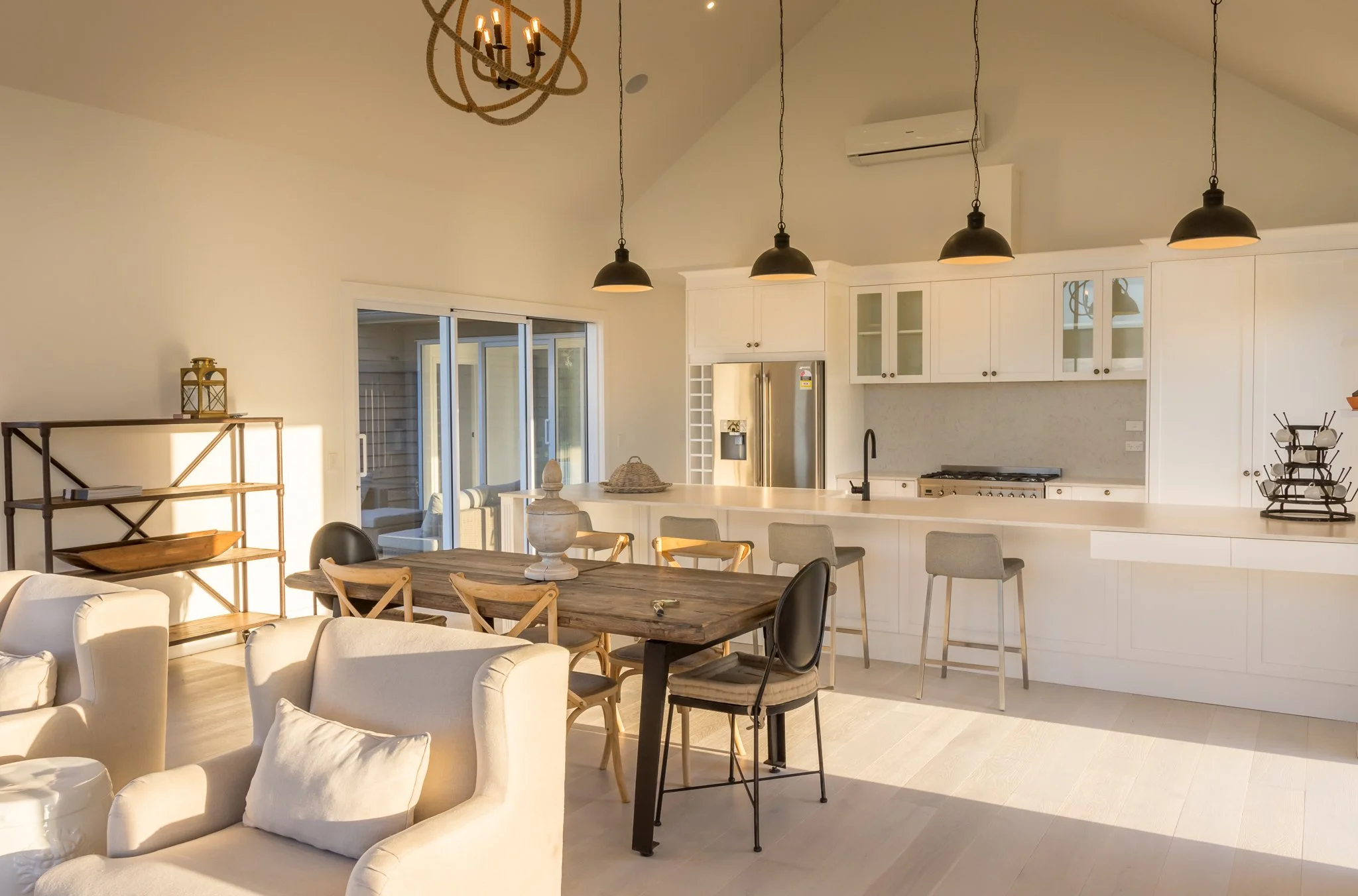 A modern open-concept kitchen and living room area with white cabinets, a stainless steel refrigerator, and black pendant lights hanging from the ceiling. There is a wooden dining table surrounded by chairs, a bookshelf, and sliding glass doors leadi