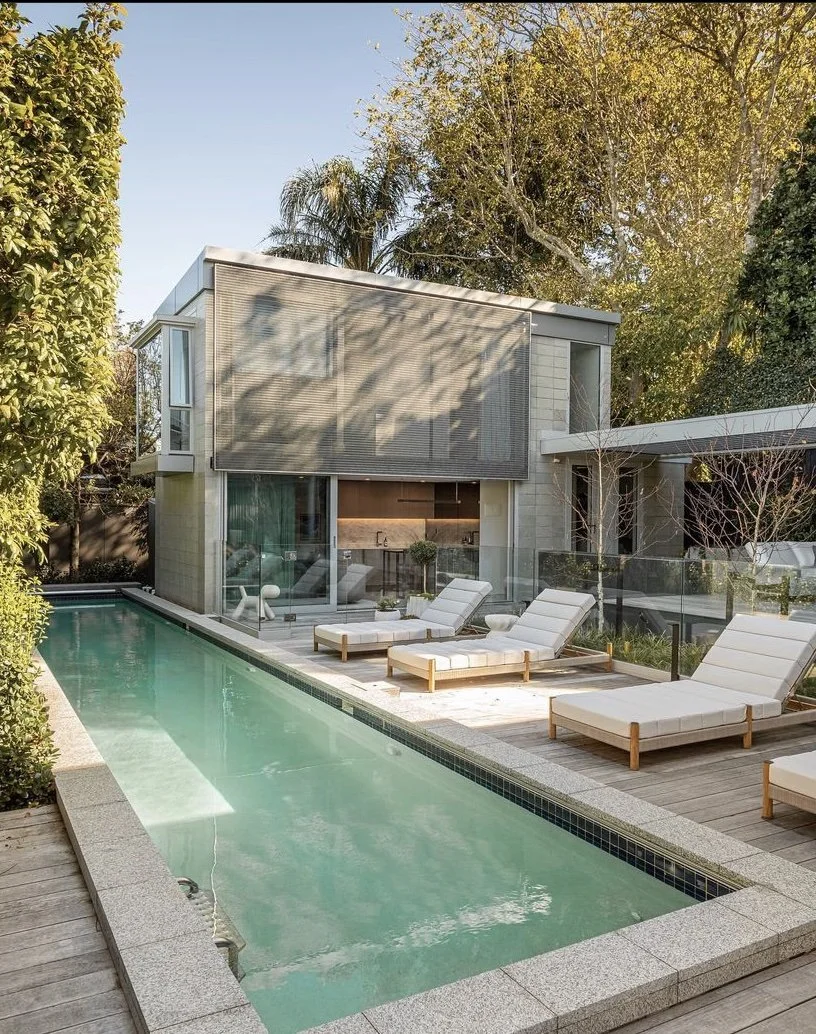 Picturesque custom modern house with a backyard pool surrounded by wooden decking and lounge chairs, trees, and a glass fence.