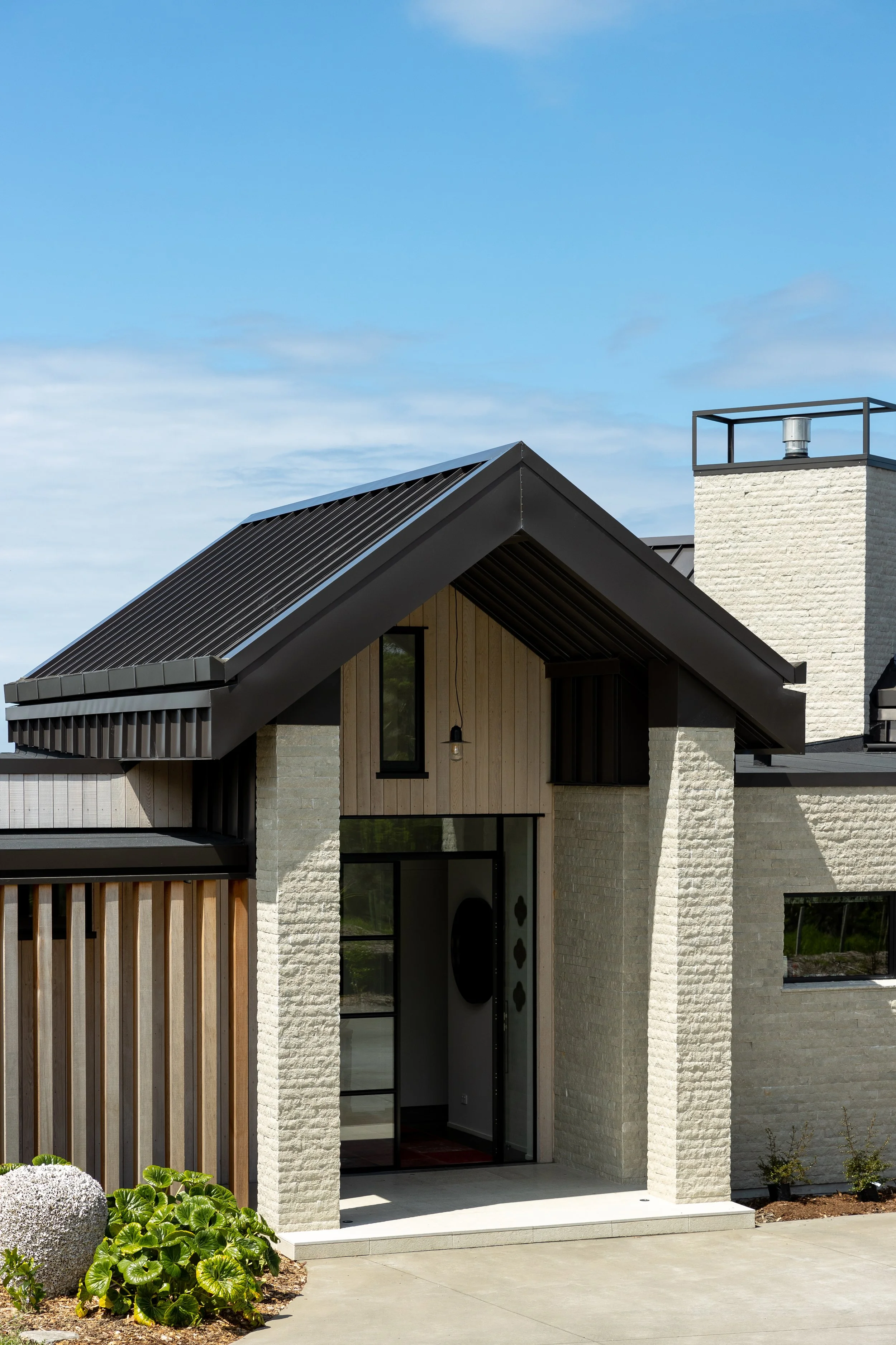 Modern house with a black sloped roof, large glass door, white brick walls, and minimalist landscaping including green plants and a gravel decorative element.