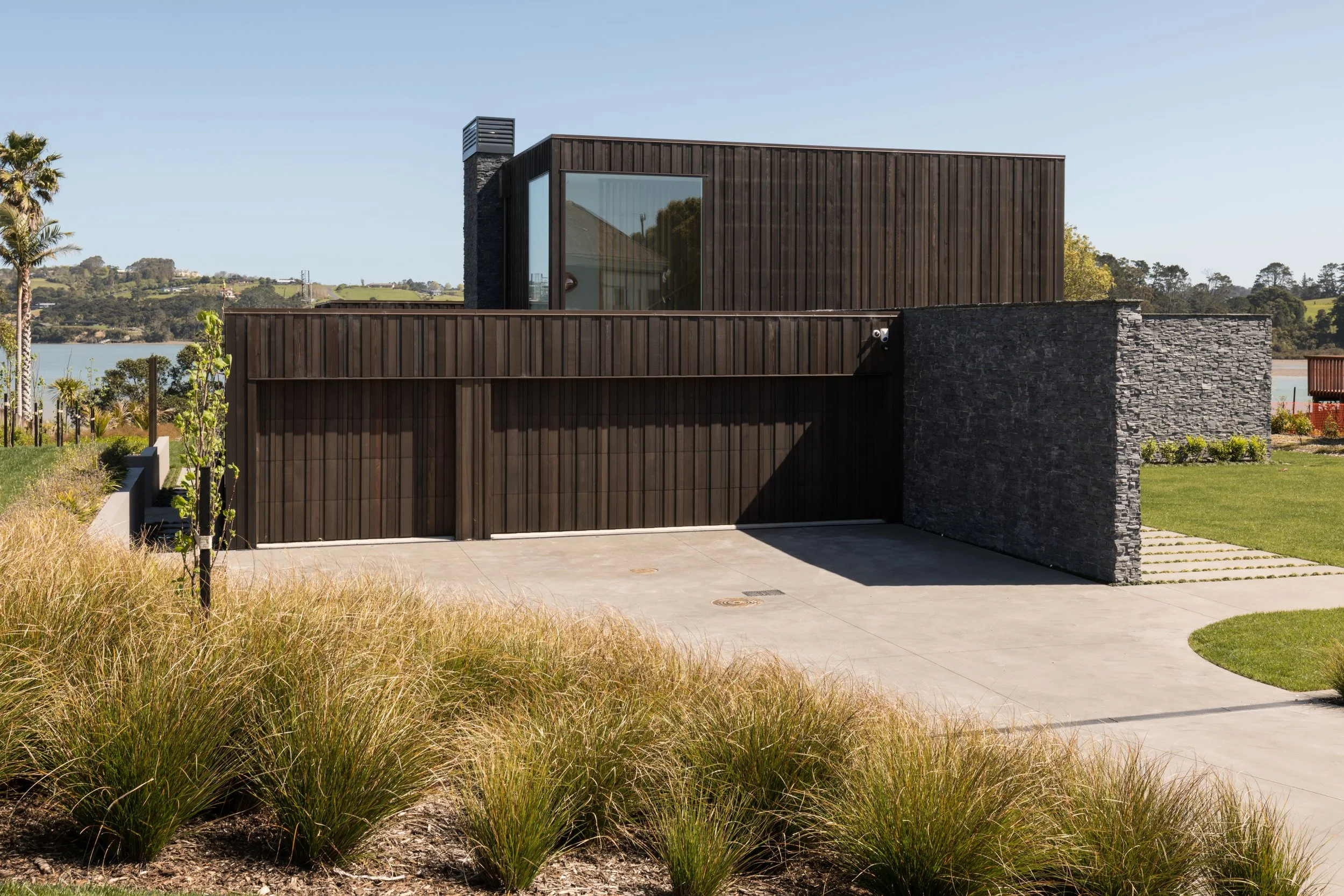 Custom modern house with dark wooden and stone exterior, large glass window, overlooking a landscape with water, trees, and hills.