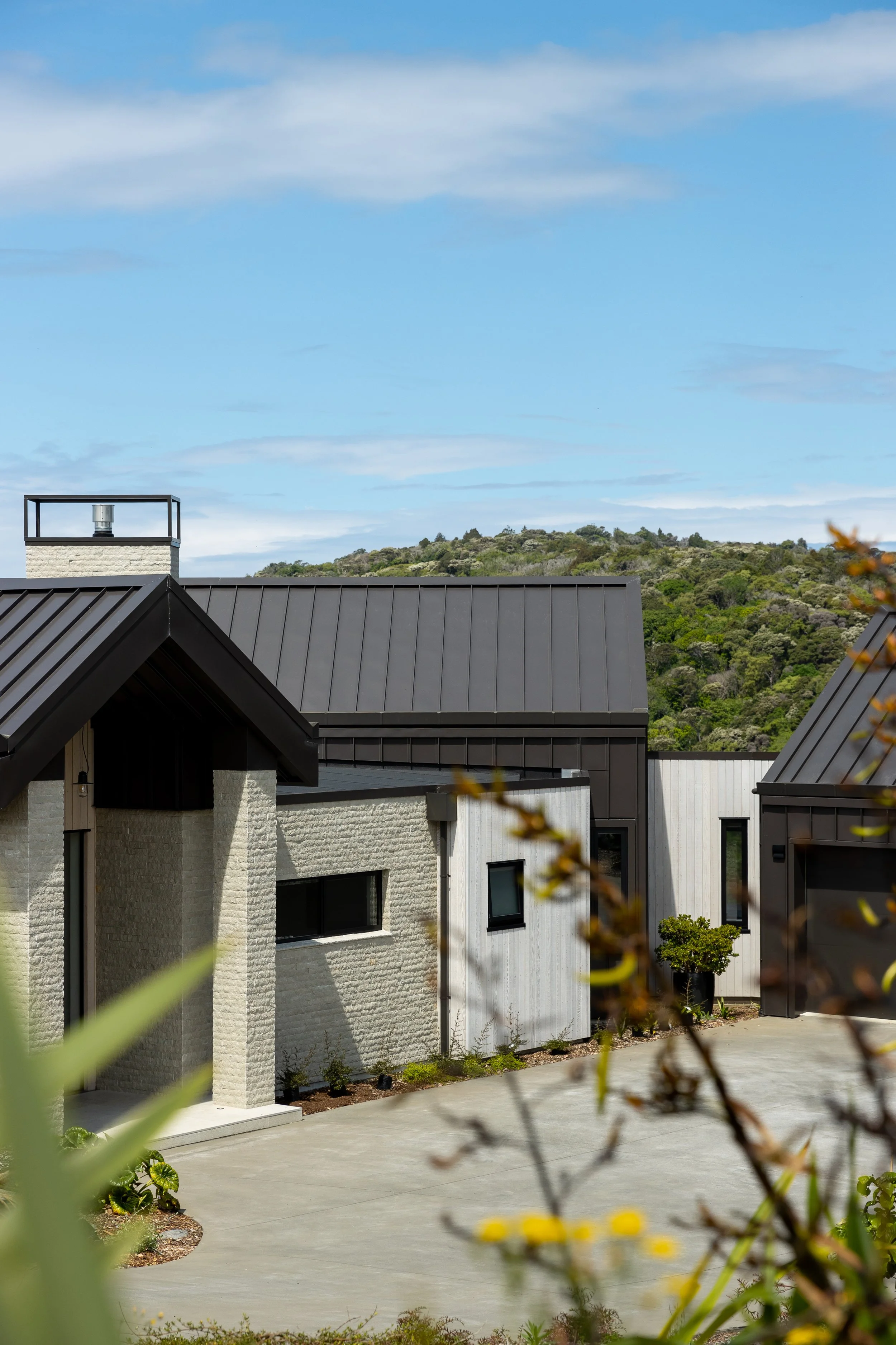 Modern residential homes with black metal roofs, beige brick walls, and large windows, surrounded by greenery and hills in the background under a partly cloudy sky.