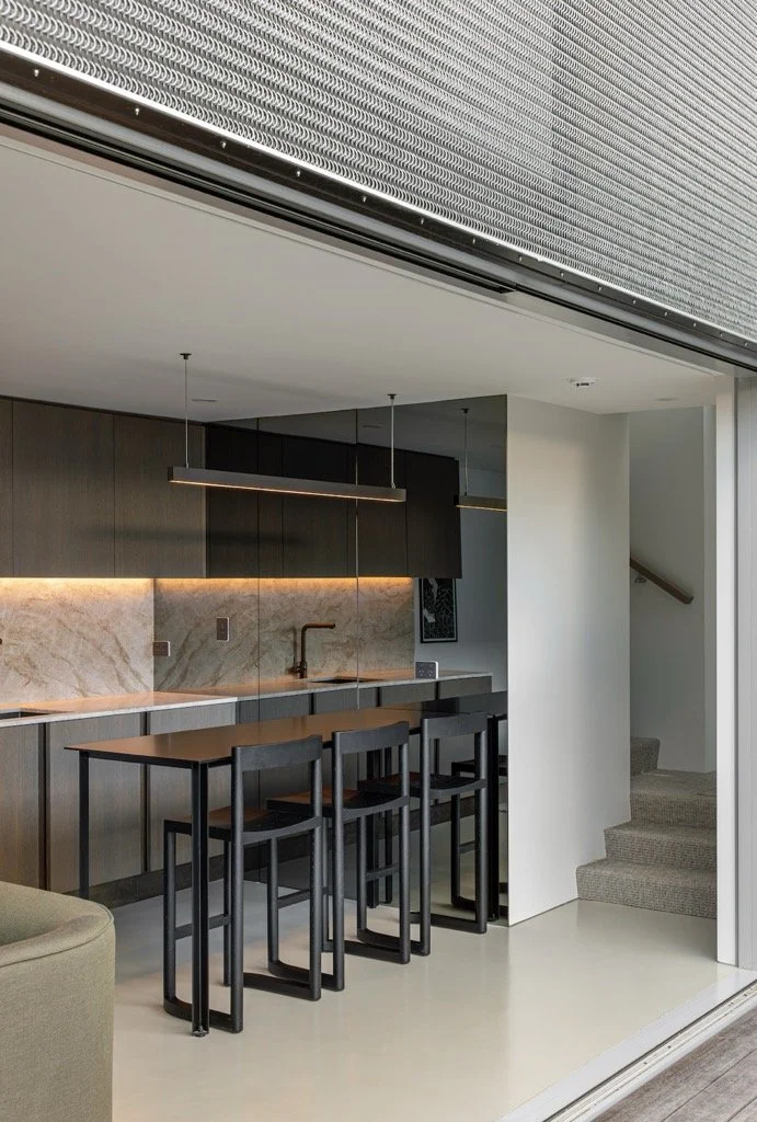 Custom modern kitchen with dark wood cabinets, marble backsplash, black high chairs, and minimalist lighting, adjacent to a staircase with gray carpet.