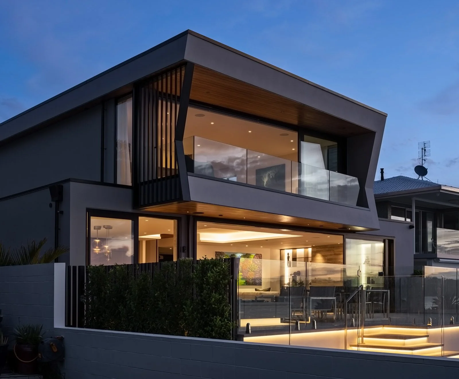 Modern luxurious custom house with large glass windows and balcony looking out to the ocean, illuminated at dusk, featuring sleek architectural angular design with wood and metal accents.