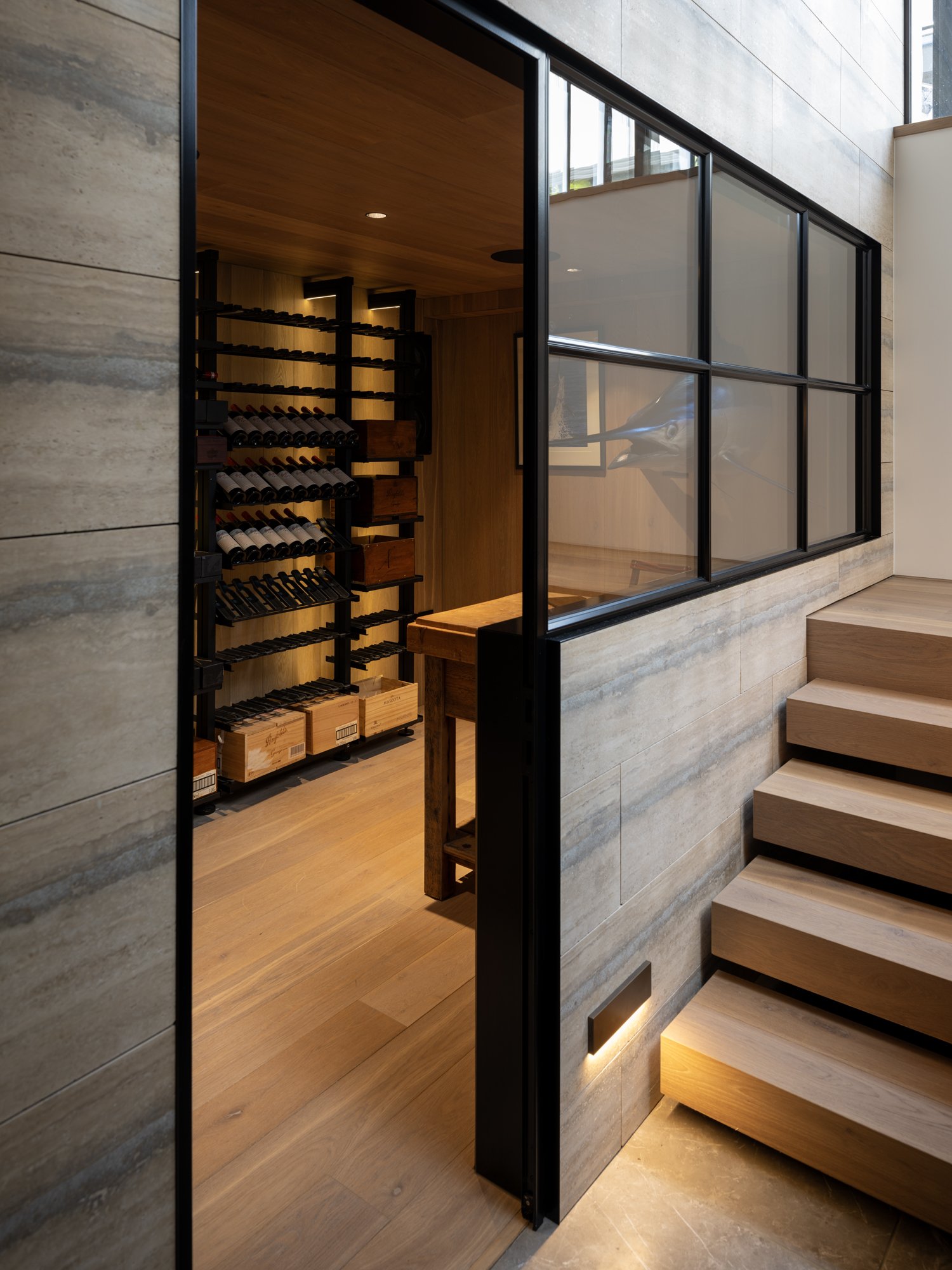 Interior of a modern architectural newbuild home with a custom floating staircase, a cellar filled with wine bottles, cedarwood paneling, and large glass windows.