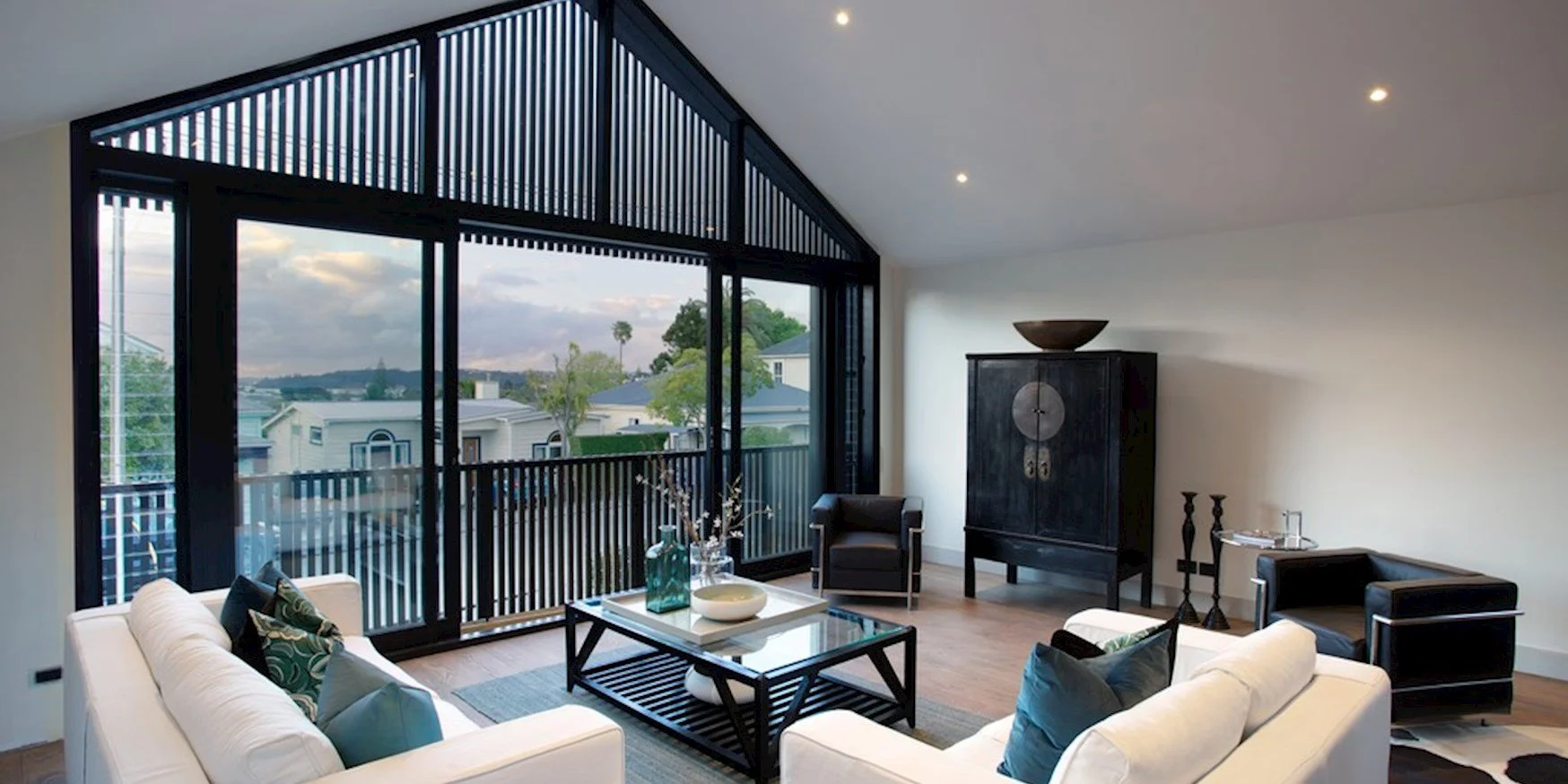 Modern living room with large windows overlooking a neighborhood and trees, white sofas with pillows, a black coffee table with decor, black armchairs, a black cabinet with a round bowl on top, and decorative candle holders.