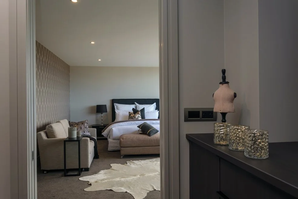View into a modern bedroom with a bed, bench, sofa, and nightstands, seen through an open door. Decorative pillows and lamps are on the bed, and a patterned accent wall is on one side.