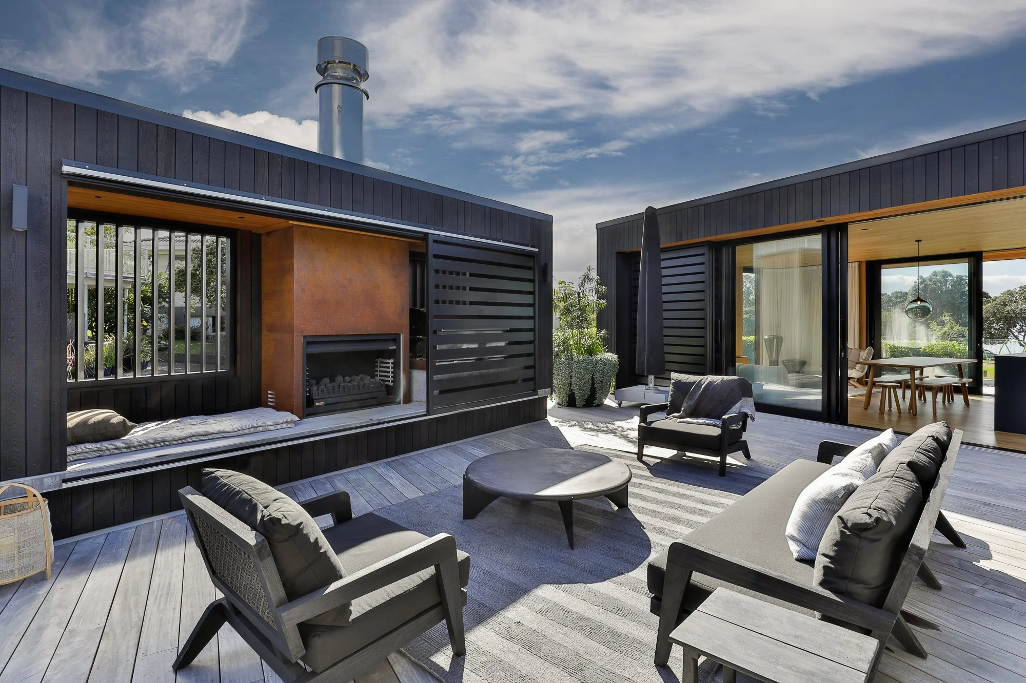 Modern outdoor patio with black wooden house facade, seating area with chairs and sofas, round coffee tables, large sliding glass doors with view into a room, and a built-in fireplace, under a partly cloudy sky.