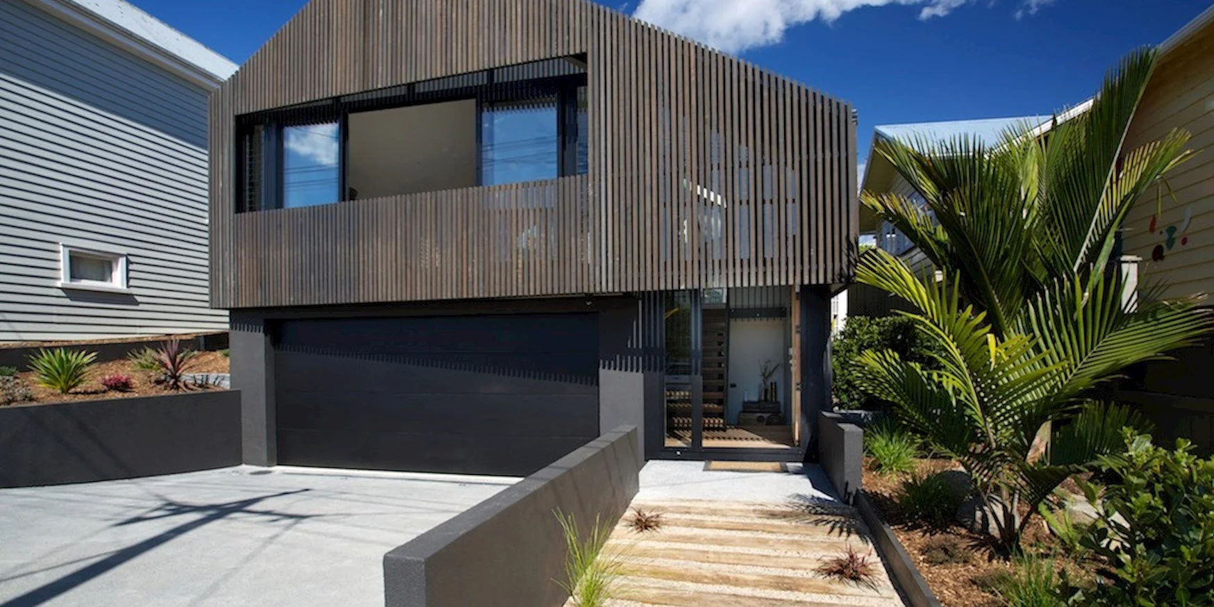 Modern two-story house with a brown wooden exterior on the upper level, a black garage door on the ground level, and landscaping with palm plants and other greenery in front.
