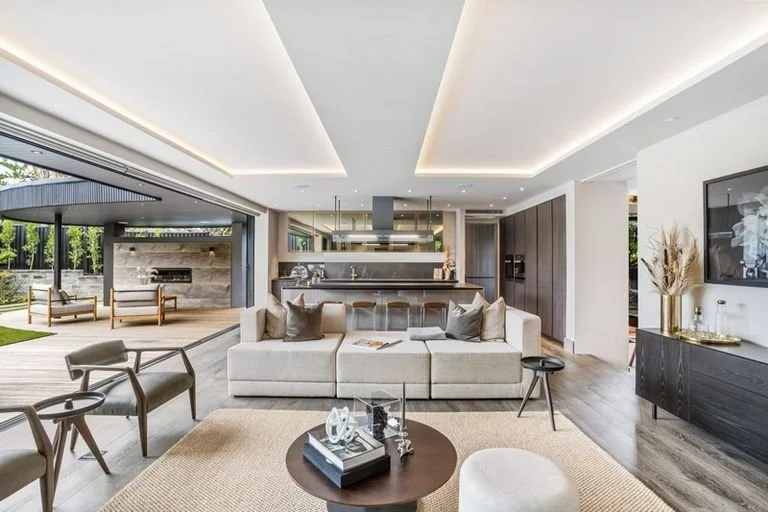 Modern open-concept living area with neutral-colored furniture, a large sofa, armchairs, a rug, and a view of an outdoor patio with chairs and a fireplace.