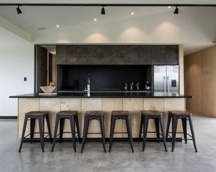 Modern kitchen with black countertop, beige lower cabinets, dark upper wall panel, stainless steel refrigerator, and five wooden bar stools at the breakfast bar.
