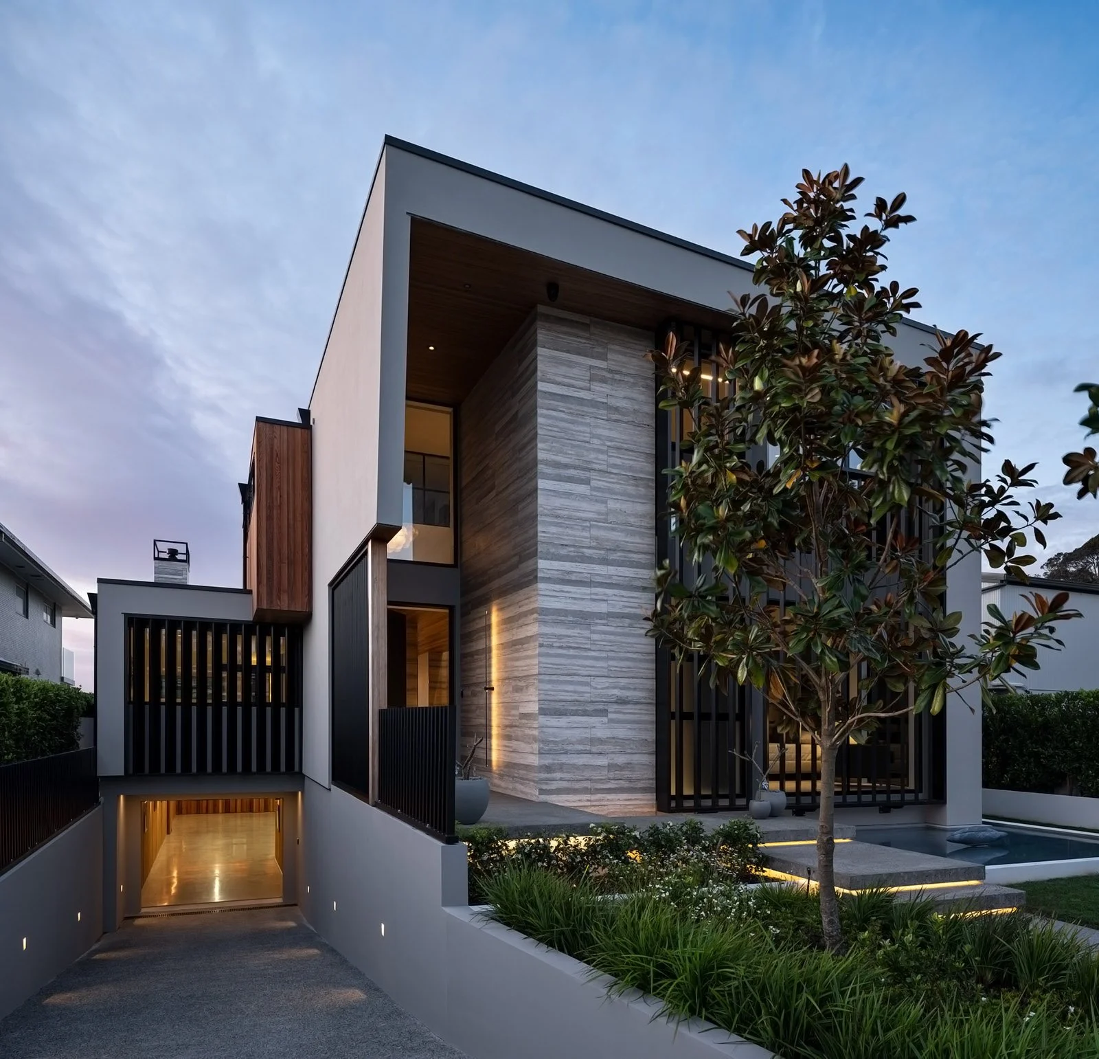 Modern multi-story house with a sleek design, featuring a mix of white, wood, and stone exterior finishes. The house has large windows, a front porch, and is surrounded by landscaped greenery and trees. The driveway leads to an underground garage.