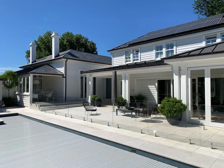 Modern white house with large windows and solar panels, adjacent to a swimming pool with a glass fence, outdoor seating, and potted plants, under a clear blue sky.