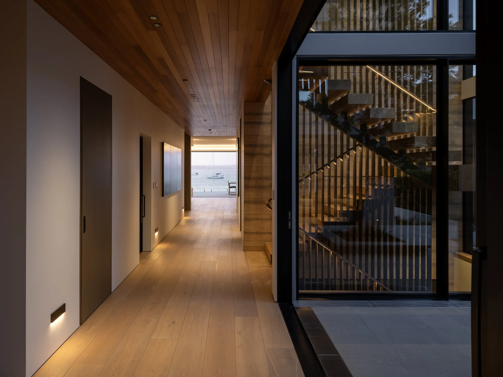 Modern indoor hallway with wooden ceiling, light-colored wooden floor, and opening showing a view of water and boats outside. To the right a feature 8.5 meter brass powder coated stairwell feature centers the home.