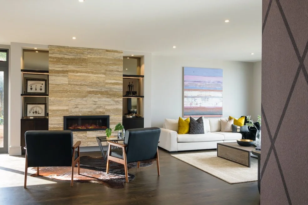 Modern living room with a fireplace, two black armchairs, a white sofa with yellow and black cushions, a painting on the wall, a rug, and a sliding glass door.