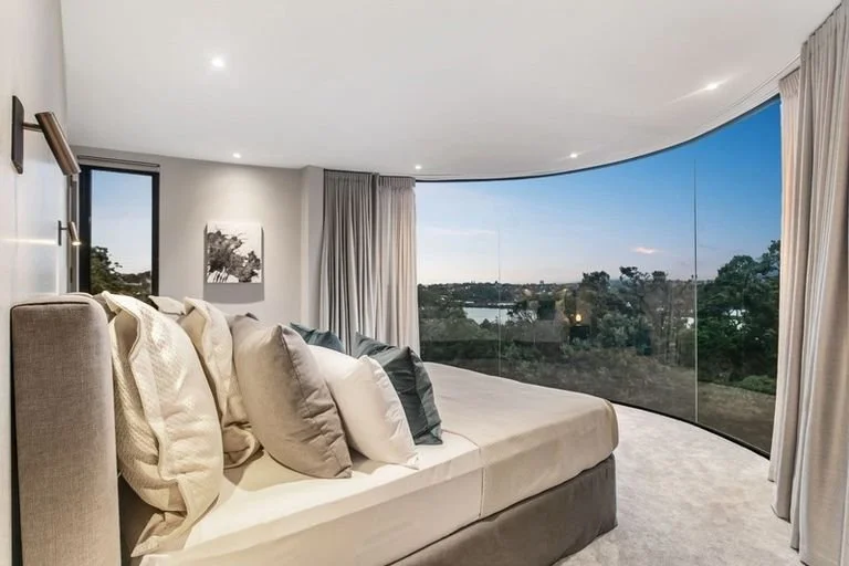 Modern bedroom featuring a large bed with multiple pillows, surrounded by beige curtains and a curved glass wall providing a view of trees and sky outside in the evening.