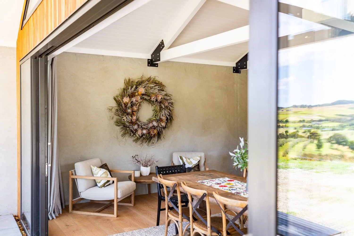 Interior of a dining area with a wooden table, chairs, and a wreath on the wall, seen through a partially open sliding glass door with curtains, overlooking a scenic landscape outside.