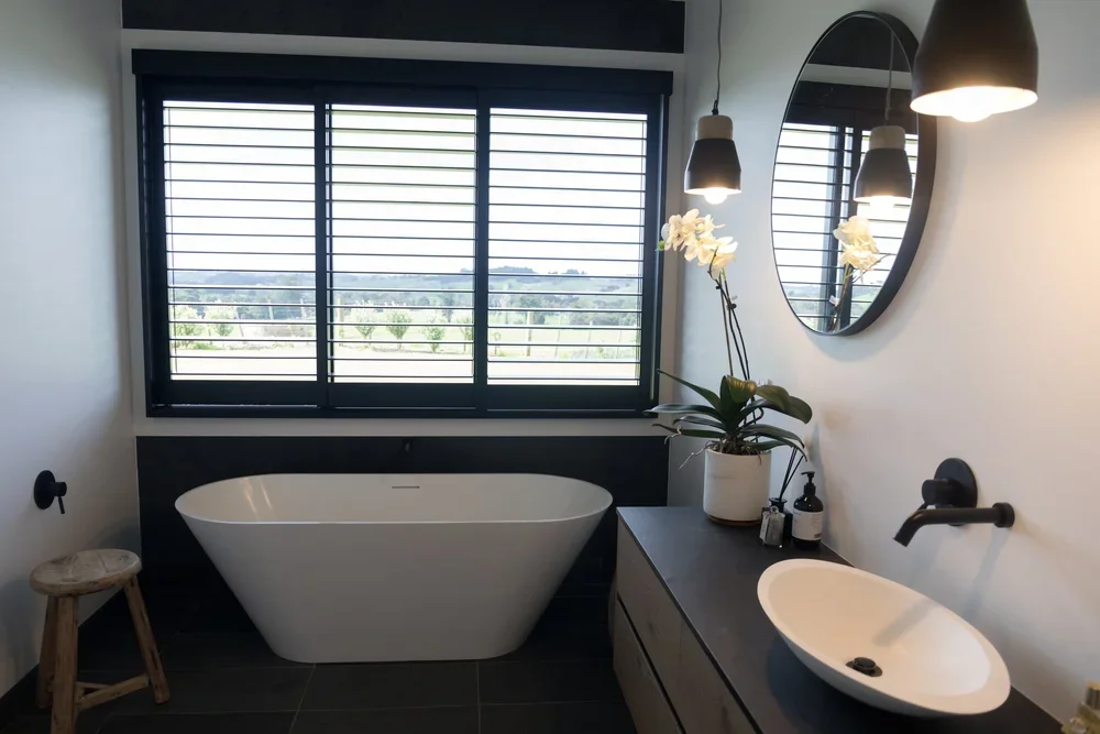 Modern bathroom with a black and white color scheme, a freestanding bathtub, a wooden stool, a potted orchid on the vanity, a round mirror, black pendant lights, and black fixtures.