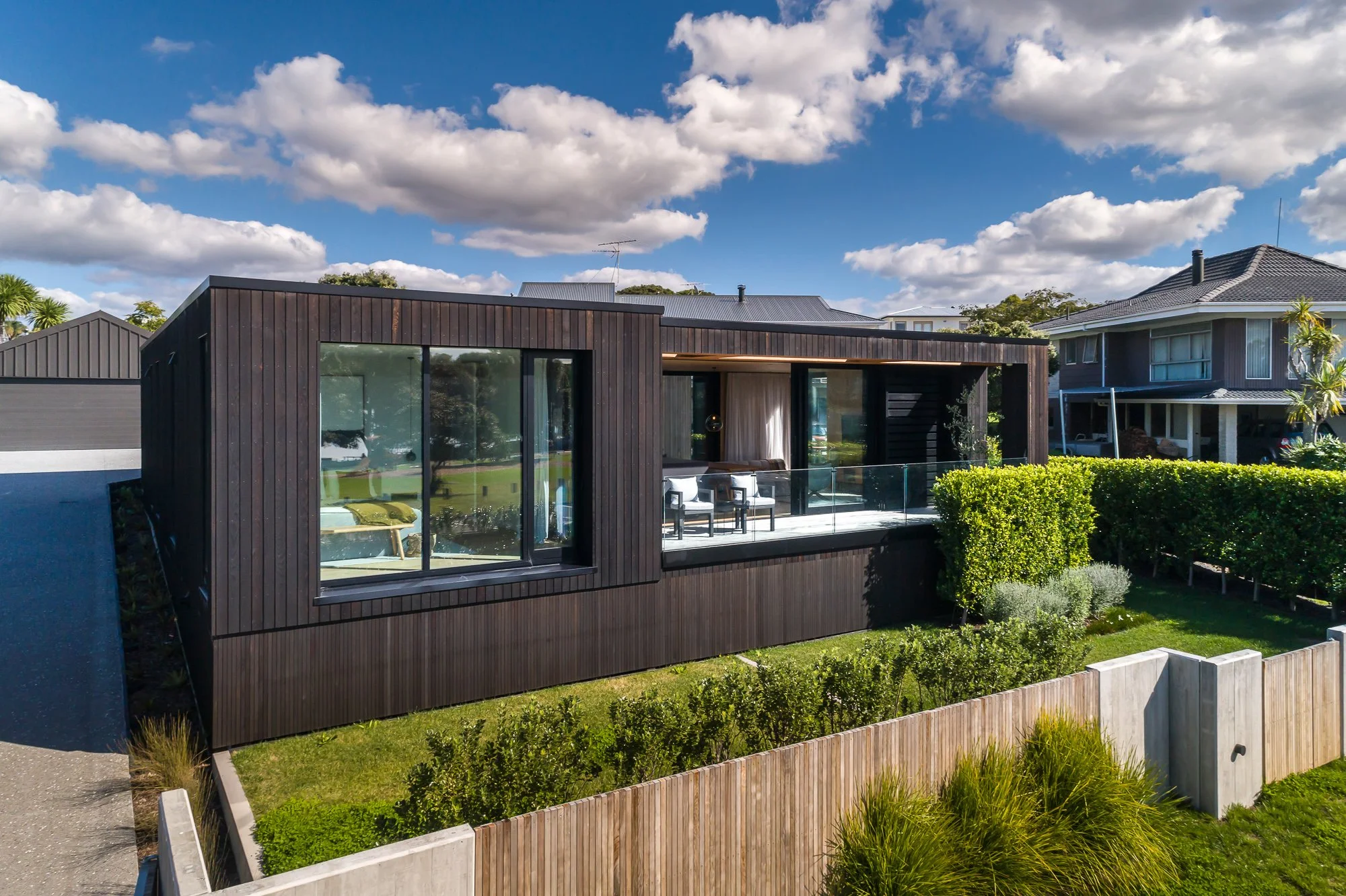 Modern black wooden cladding custom home with floor-to-ceiling windows, small outdoor seating area on balcony, surrounded by green bushes and a landscaped yard, under a blue sky with white clouds.
