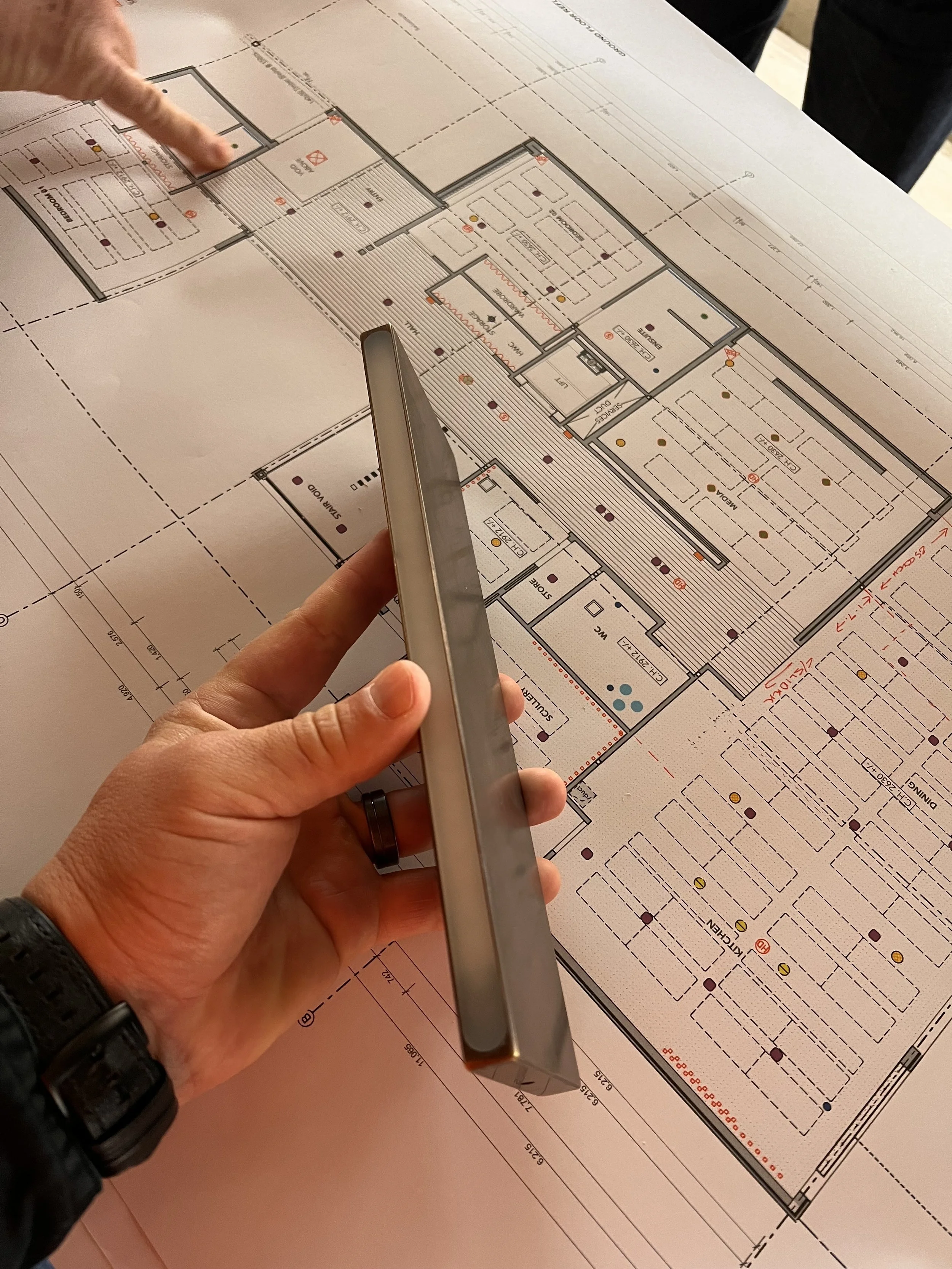 A person holding a smartphone and pointing at a large architectural floor plan on a table, with another hand visible pointing at the plan.
