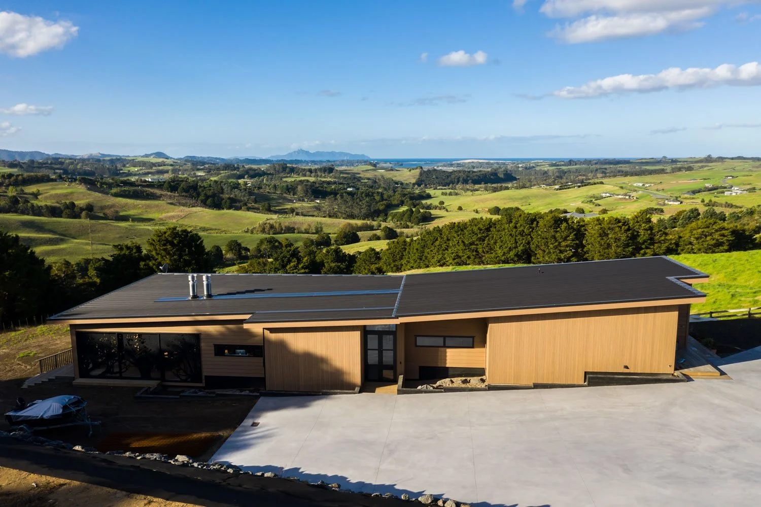 A modern single-story house with a black roof and wooden siding, set on a large concrete driveway, surrounded by green rolling hills and a partly cloudy sky.