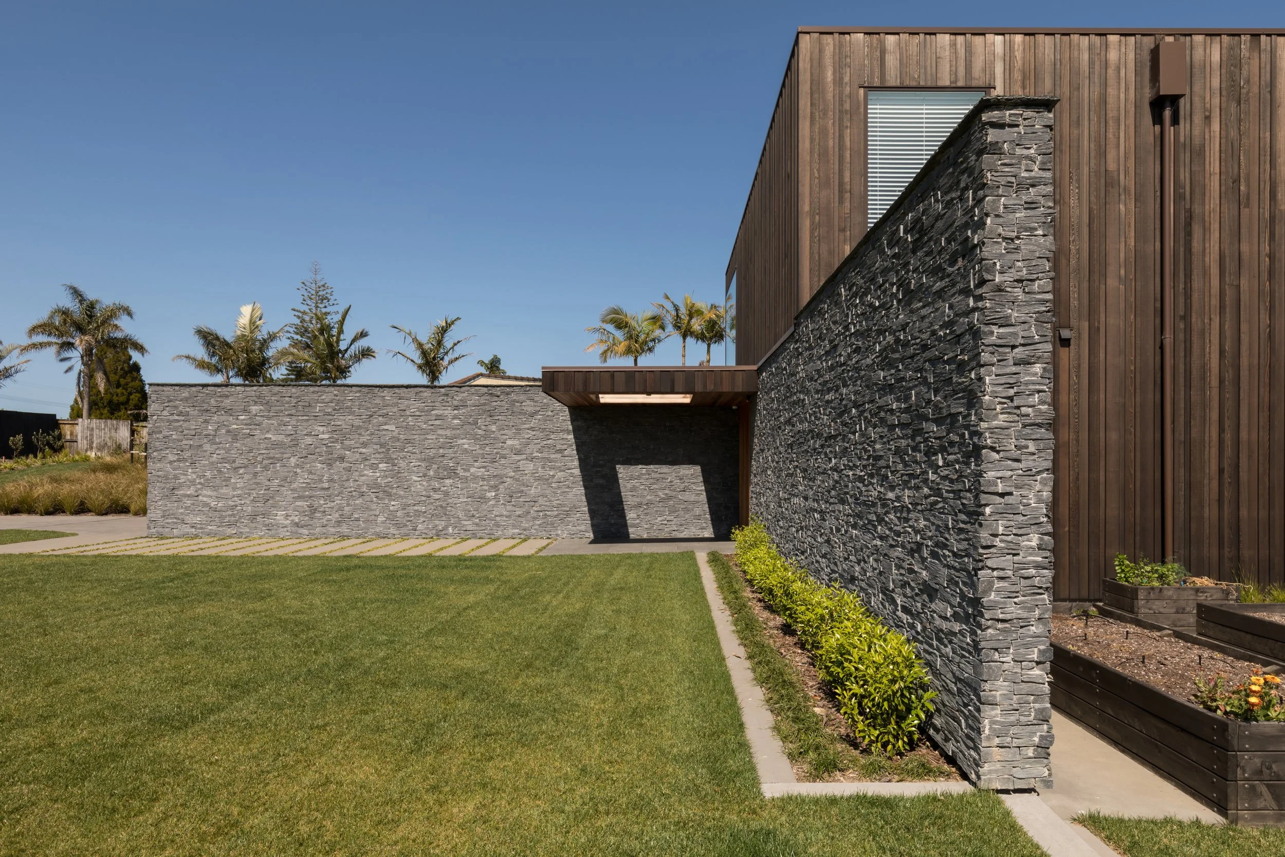 Contemporary house with a stone wall, wooden siding, and a window, surrounded by a well-maintained grassy yard and small shrubs, under a clear blue sky with palm trees in the background.