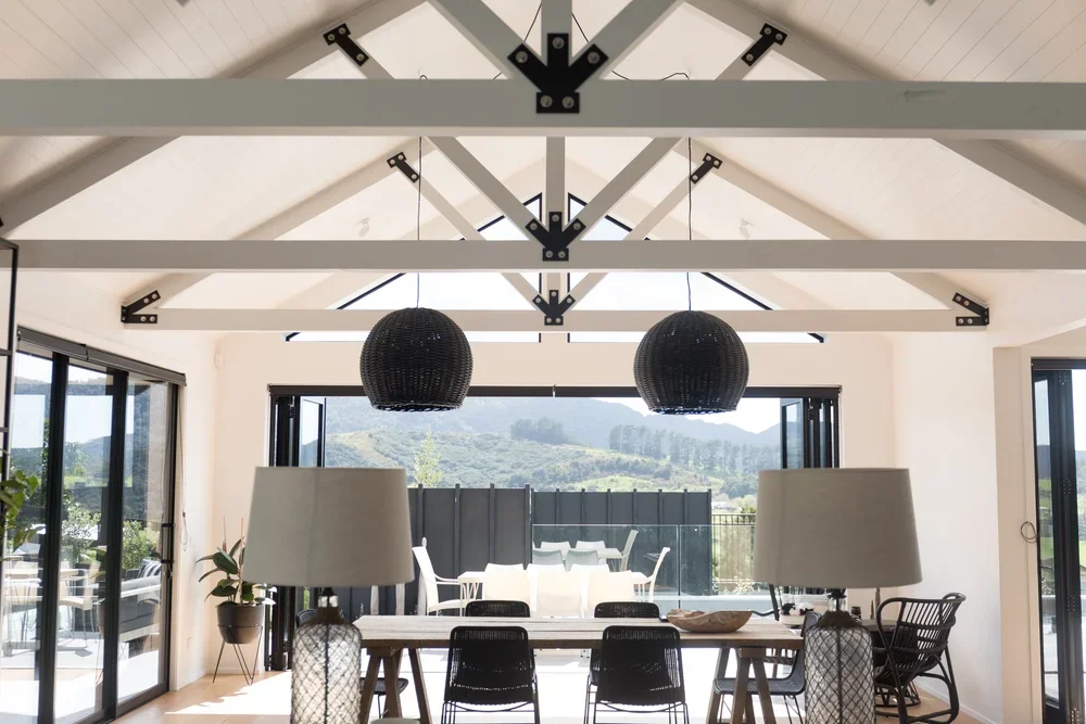 Bright dining room with large glass doors opening to a patio and mountain view, featuring a wooden dining table, black chairs, two large table lamps, and two black woven pendant lights, with white wooden beams on the ceiling.