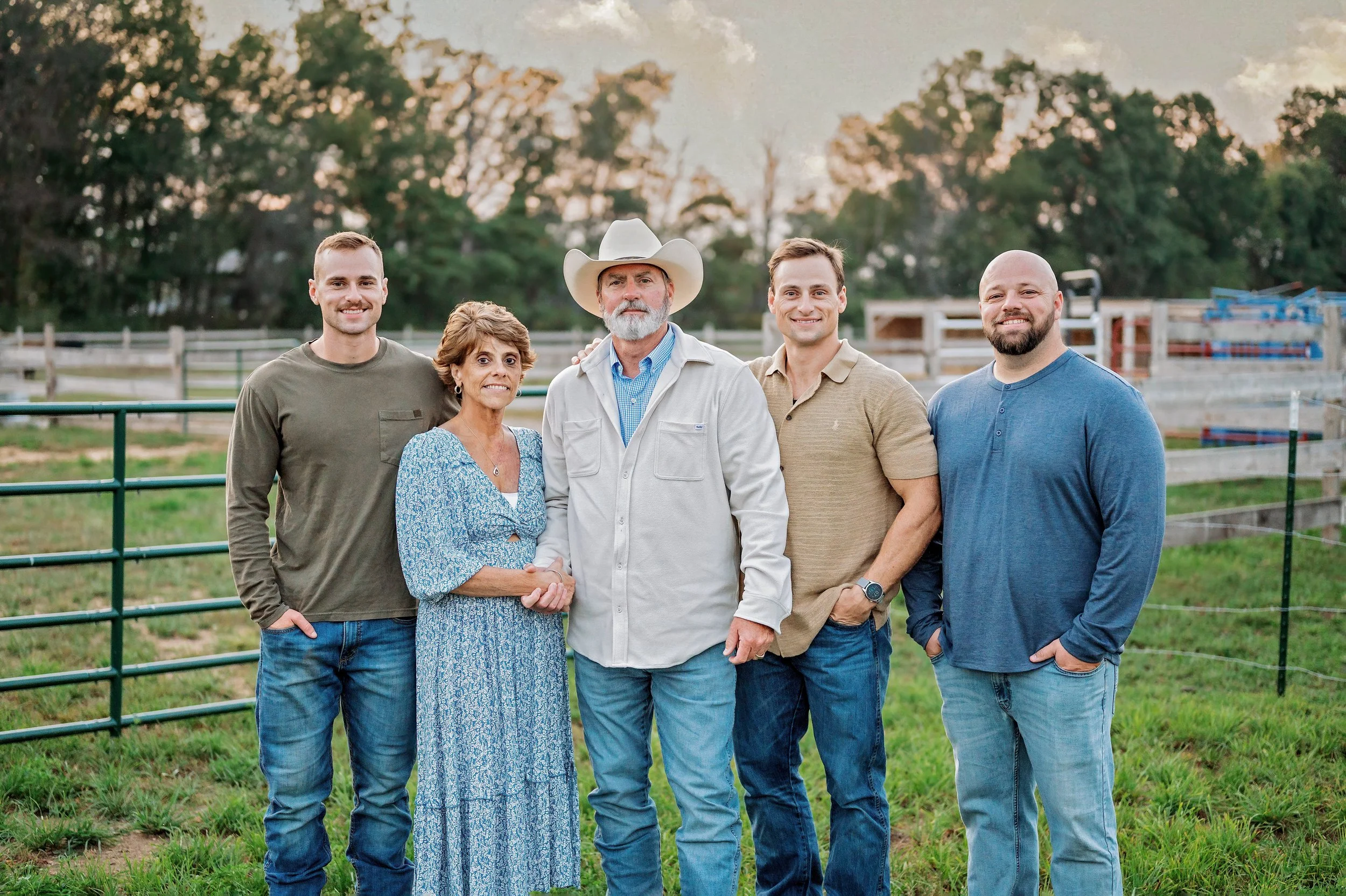 A group of six people standing together outdoors on a farm or ranch, smiling at the camera with trees and fencing in the background, during what appears to be the late afternoon or early evening.