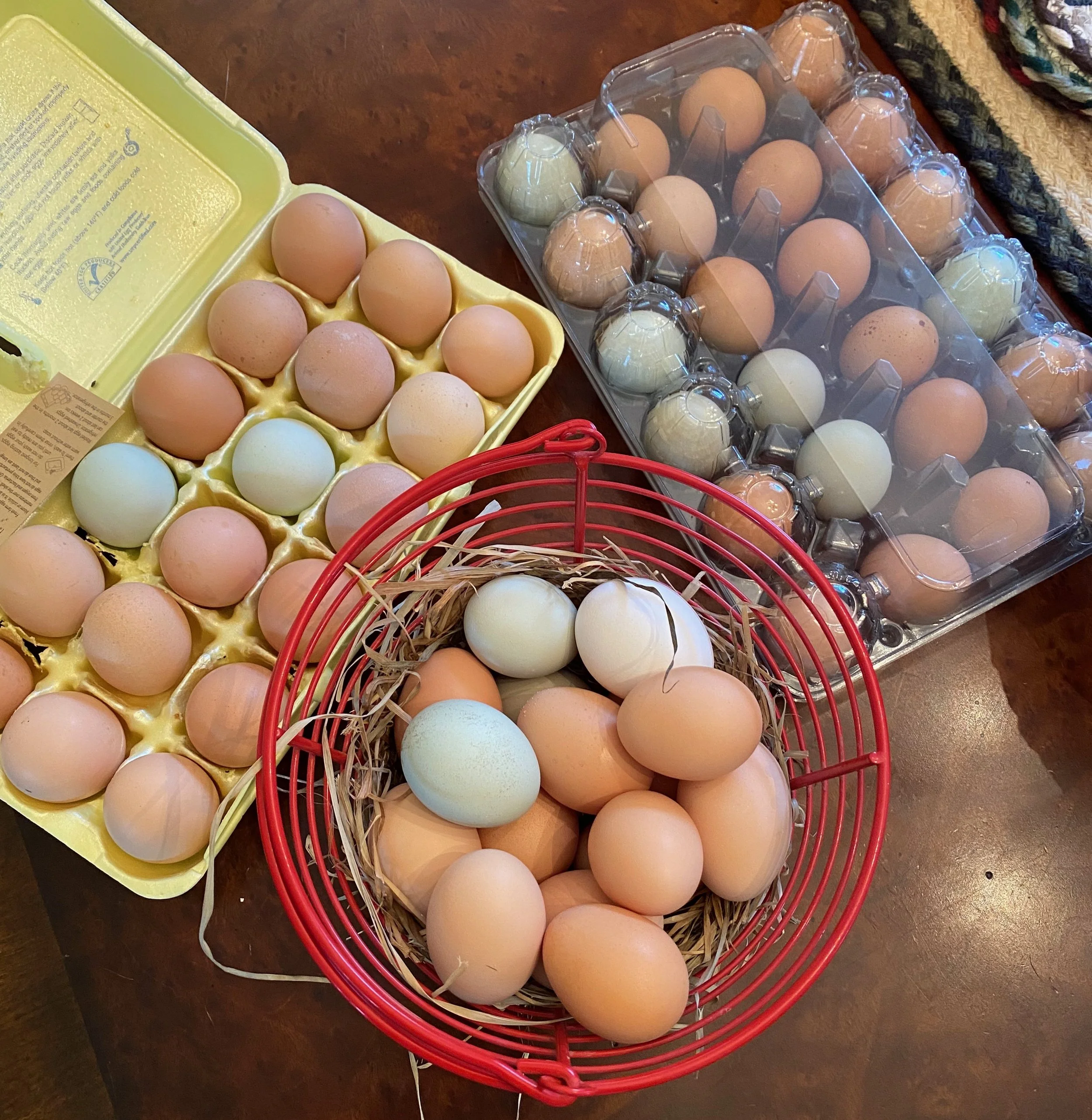 A yellow egg carton filled with brown eggs, a plastic container with more brown eggs, and a red basket with white eggs on a wooden surface.