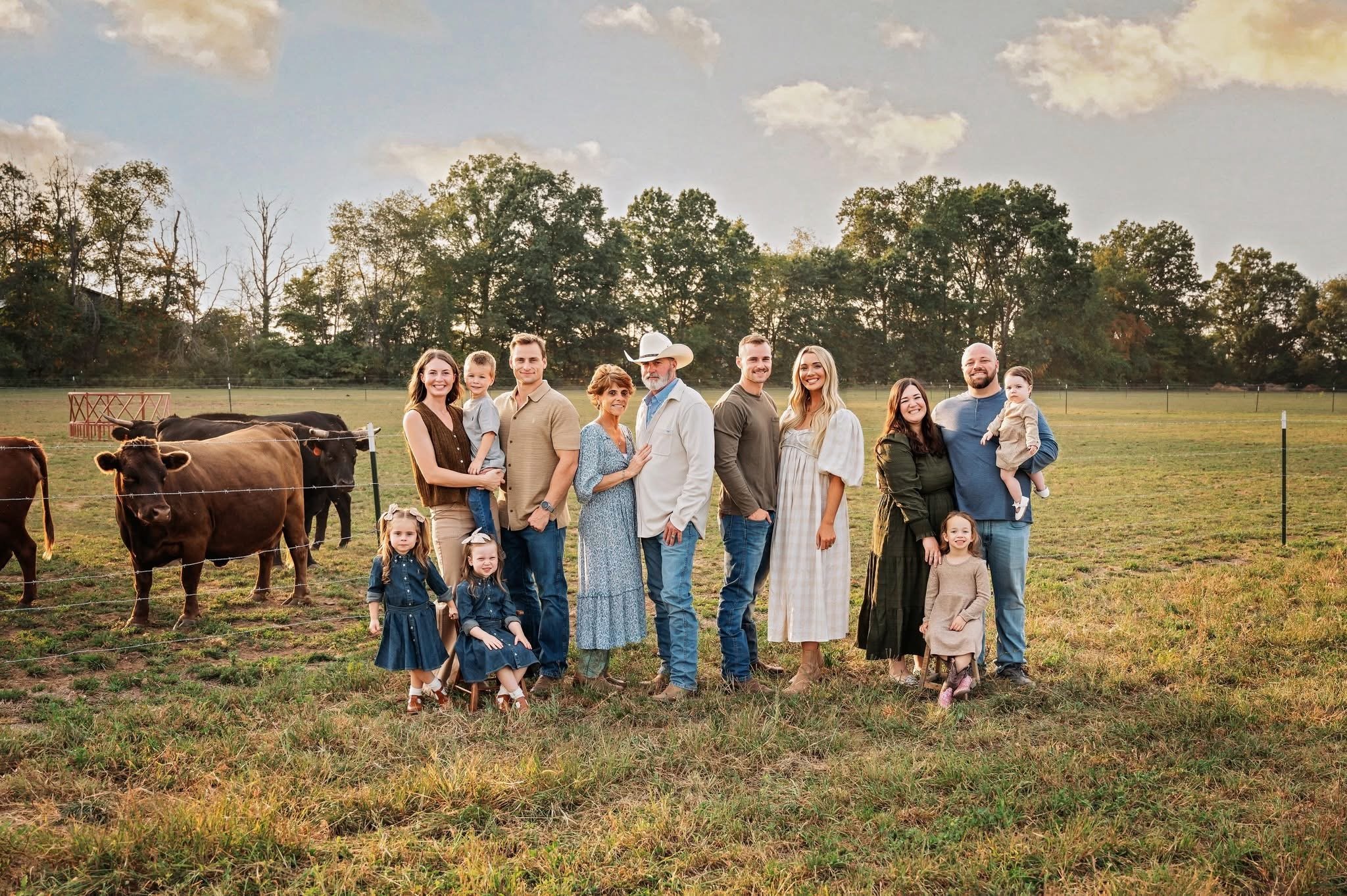A group of people, including children and adults, standing outdoors in a field with cows in the background during sunset.