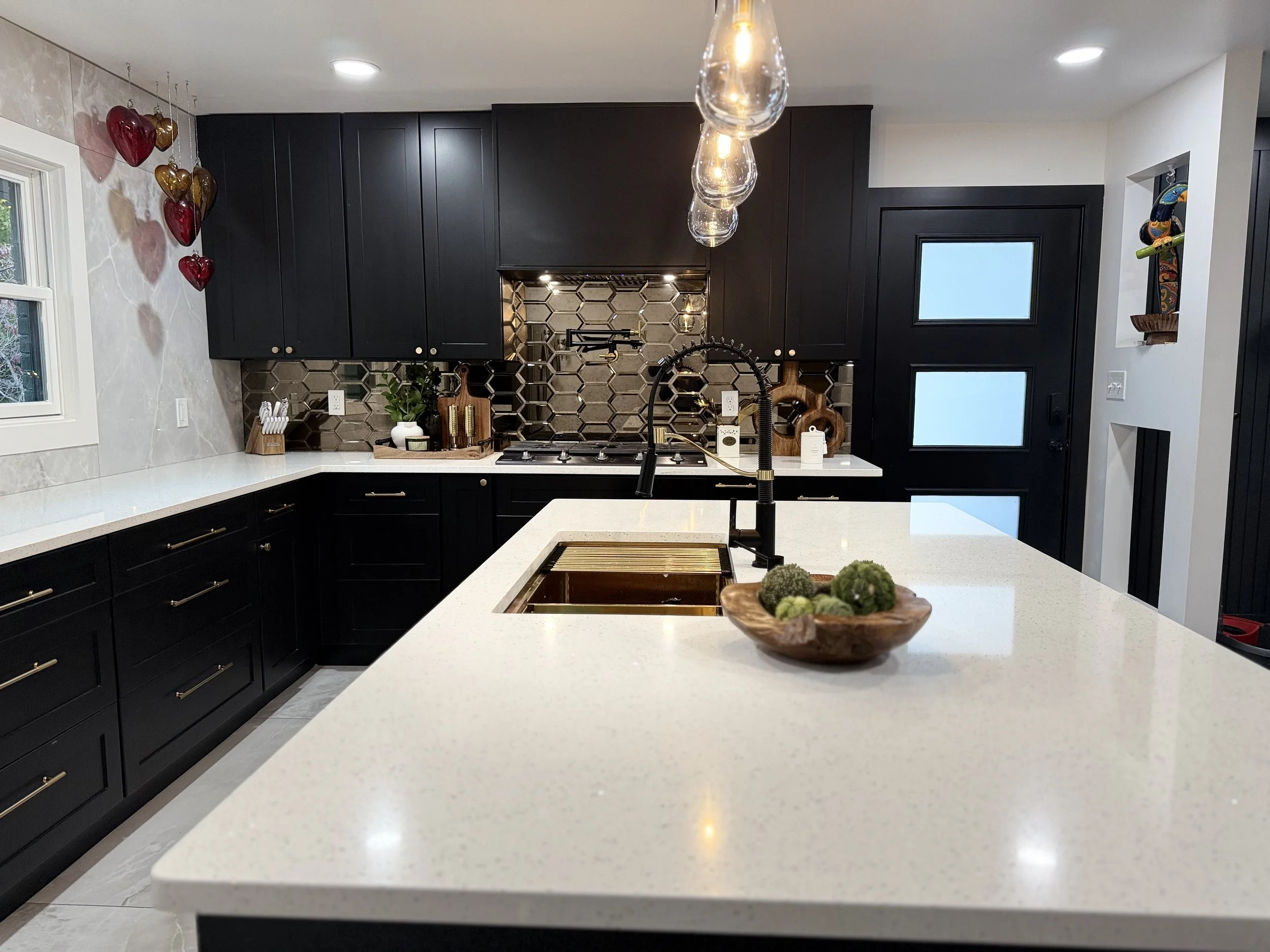 Modern kitchen with black cabinetry, white countertops, and decorative backsplash. A bowl with green decorative balls on the island, pendant lights above, and Valentine's Day heart balloons on the wall.