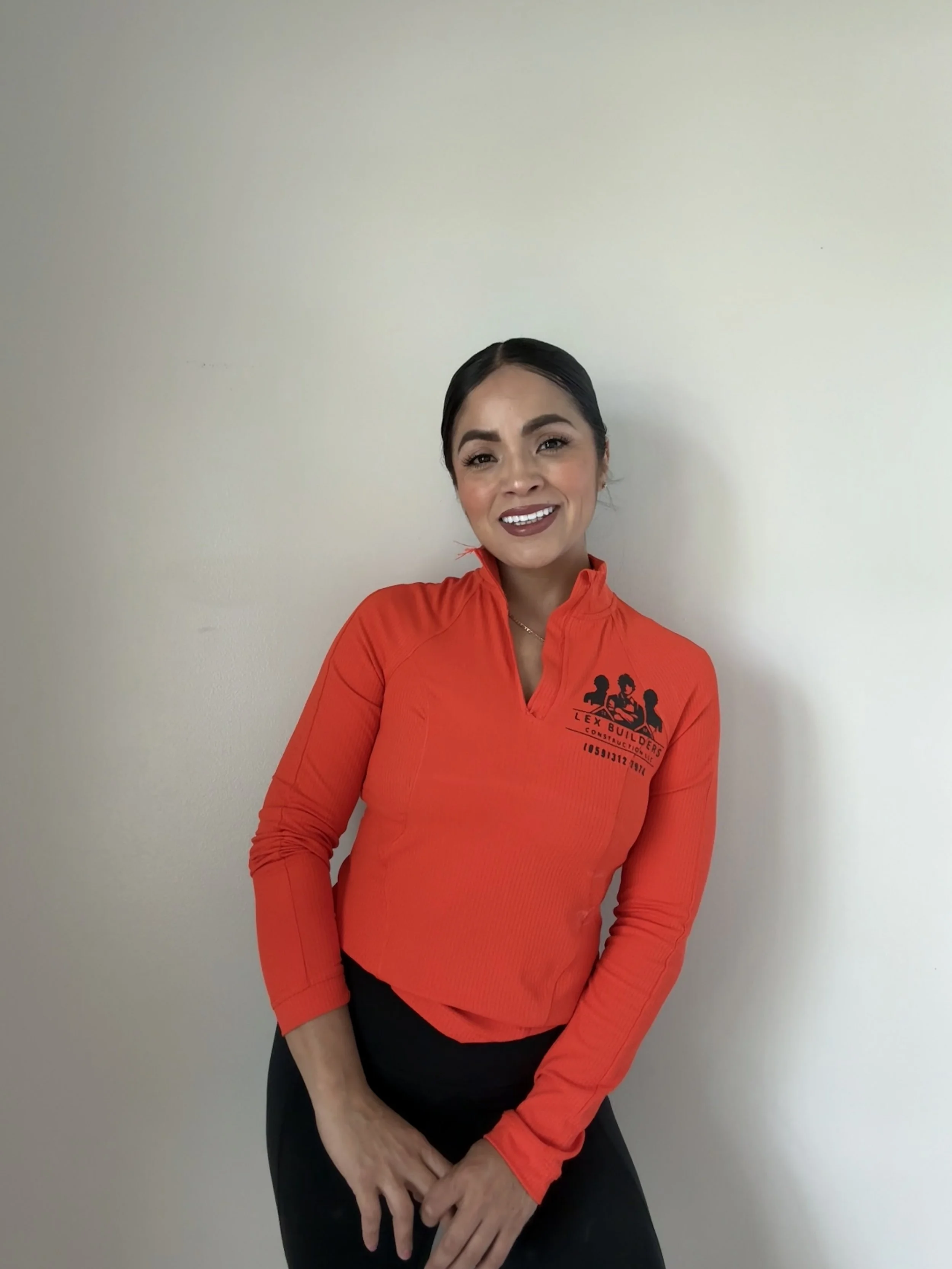 A smiling woman in a red long-sleeve athletic top with black text and graphics, standing against a plain off-white wall.