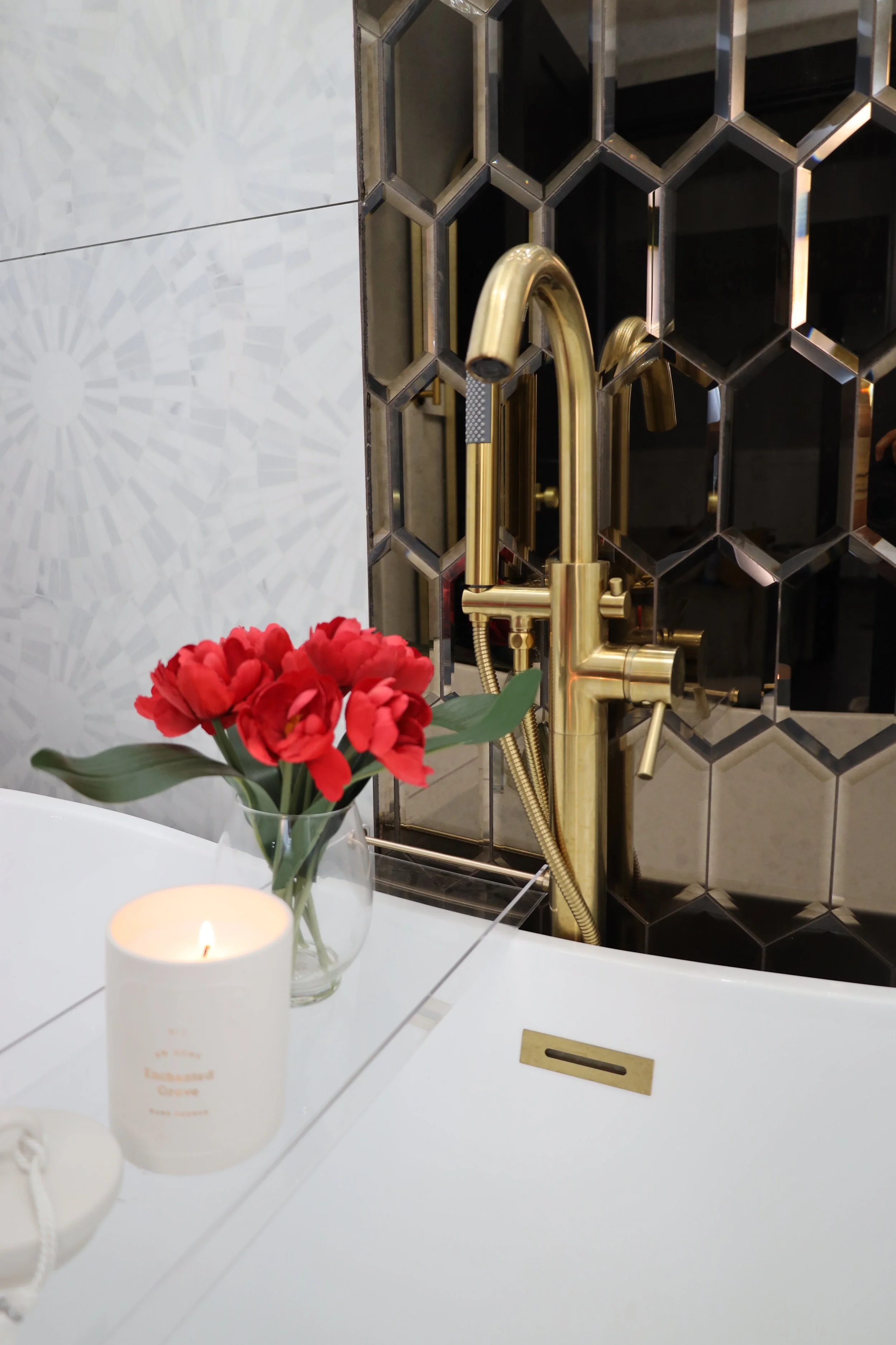 A bathroom sink area with a gold faucet, a glass vase with red flowers, a lit candle, and a white countertop.