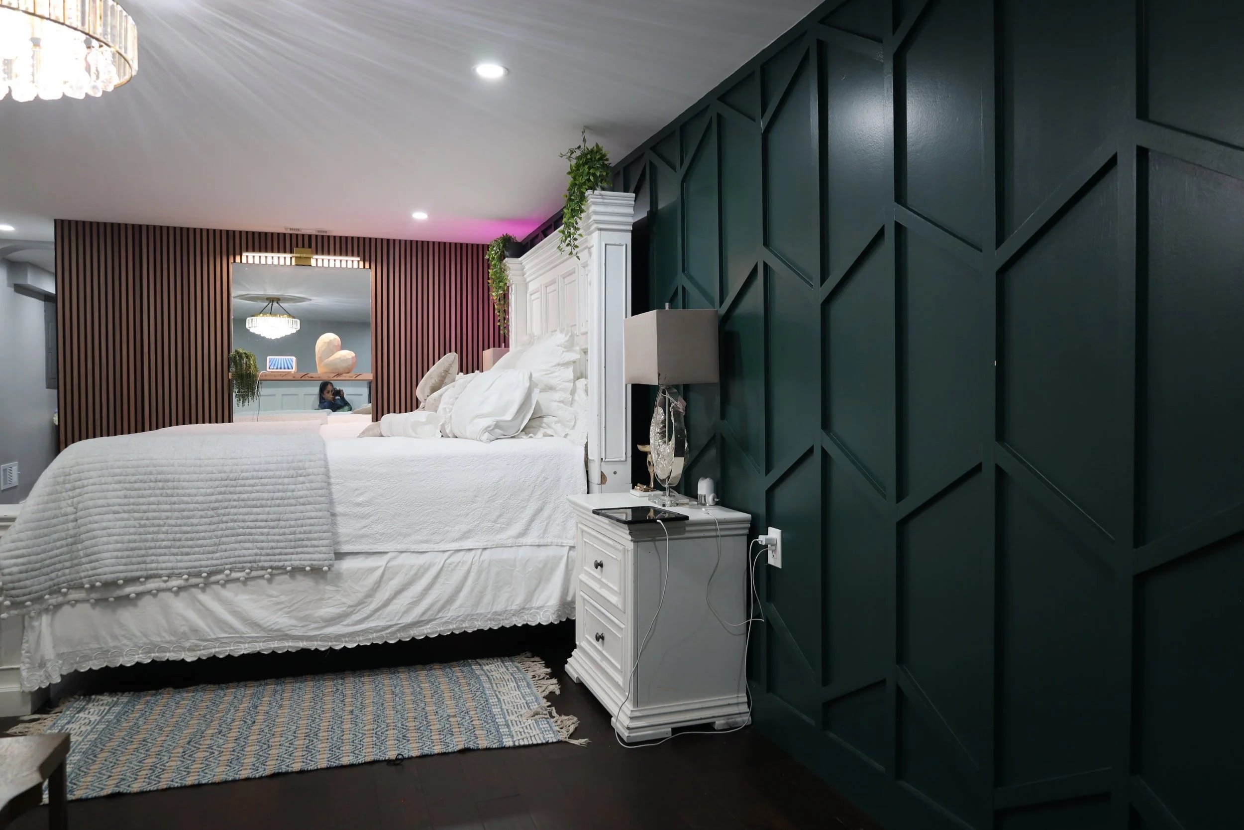 A bedroom featuring a white bed with pillows, a white nightstand, dark green geometric accent wall, and a mirror reflecting a woman sitting on a sofa.