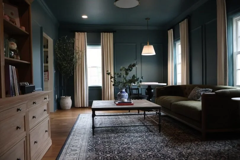 Living room with teal walls, wooden flooring, a green sofa, a coffee table with a vase and books, a large potted plant, and windows with cream curtains.