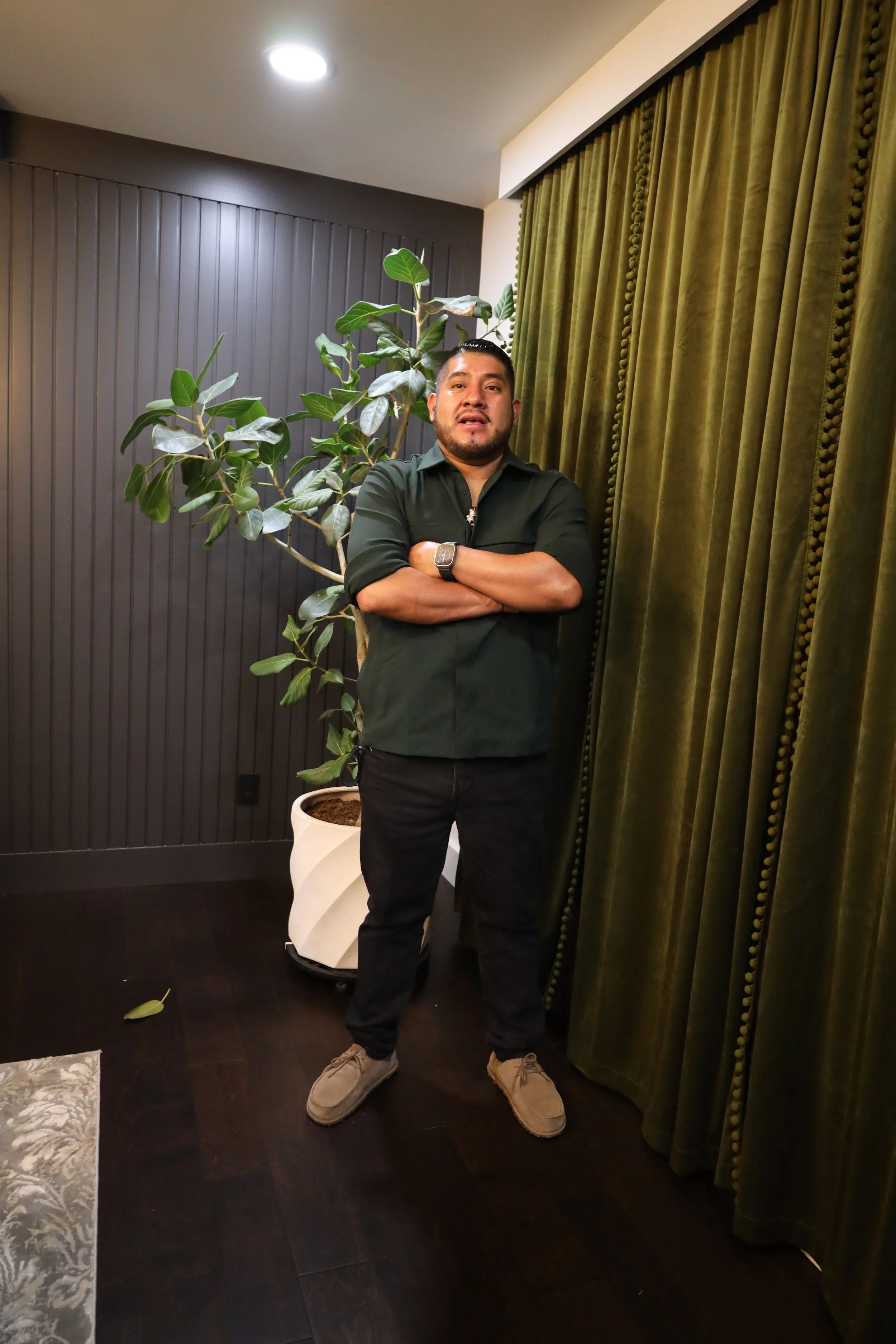 A man standing indoors with arms crossed, near a potted plant and green velvet curtains.